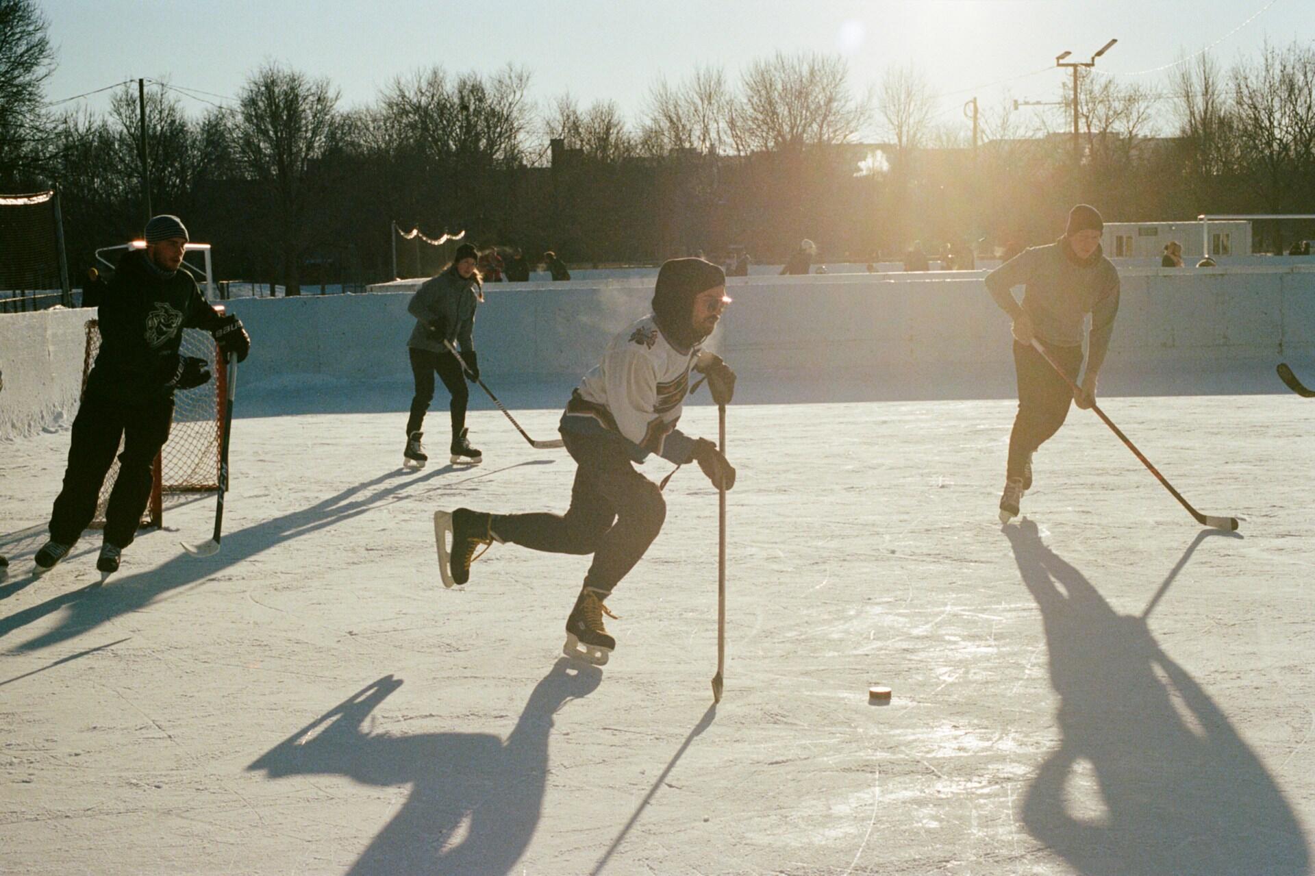 Skridskoåkare spelar hockey på en utomhusisbana, solens strålar kastar långa skuggor över isen. Träd raderar sig i bakgrunden på en solig dag.