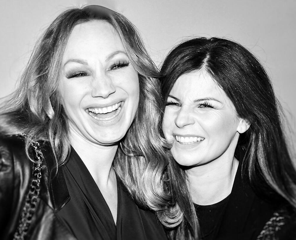 CHarlotte Perelli och Carola Häggkvist poserar tätt intill varandra, båda ler och är klädda i svarta kläder, mot en ljusgrå bakgrund.