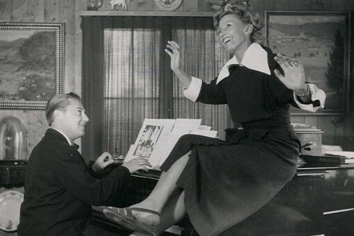 Lale Andersen sitter på ett piano och hennes make Artur Beul spelar. Bild från Wikimedia Commons. 