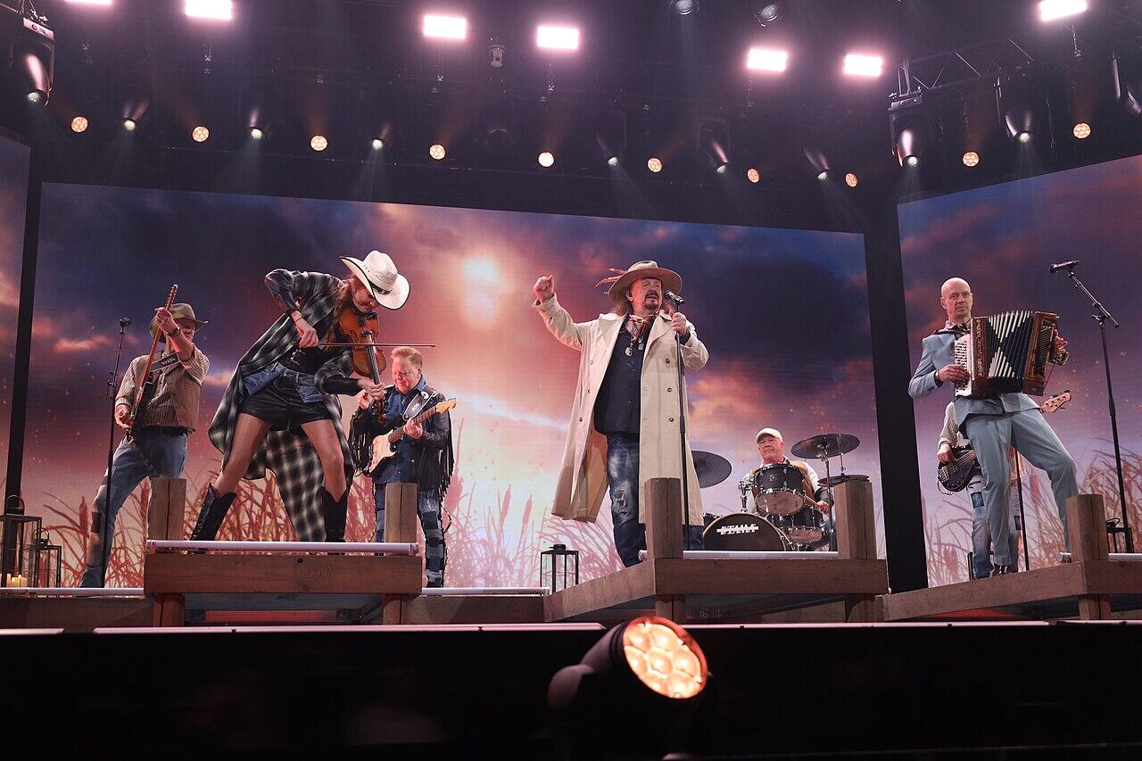 En livlig countrymusikföreställning med musiker som spelar olika instrument på scenen, mot en dramatisk bakgrund.