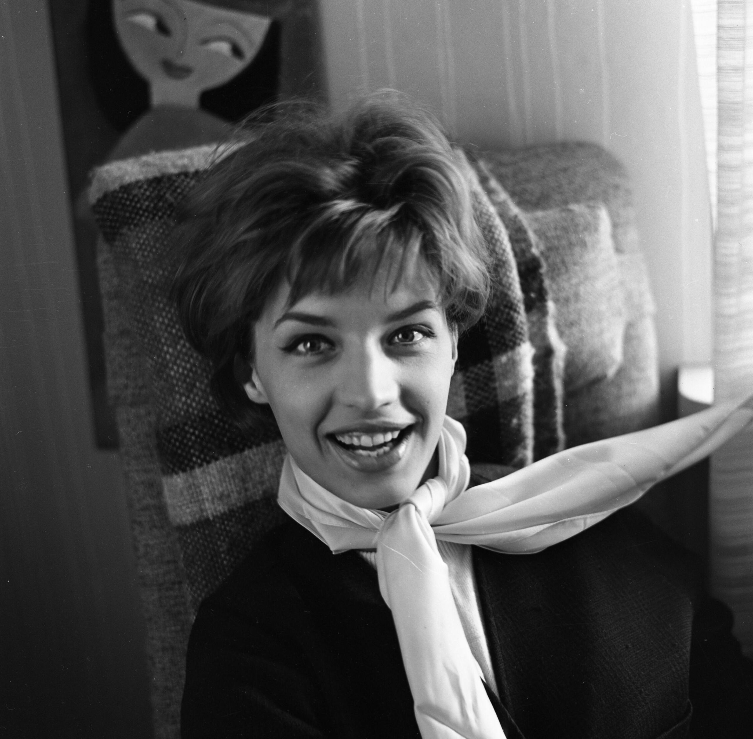 Lill Babs med lockigt hår sitter bekvämt i en mönstrad stol, bär en stilfull scarf och omges av mjuka skuggor från solljuset.