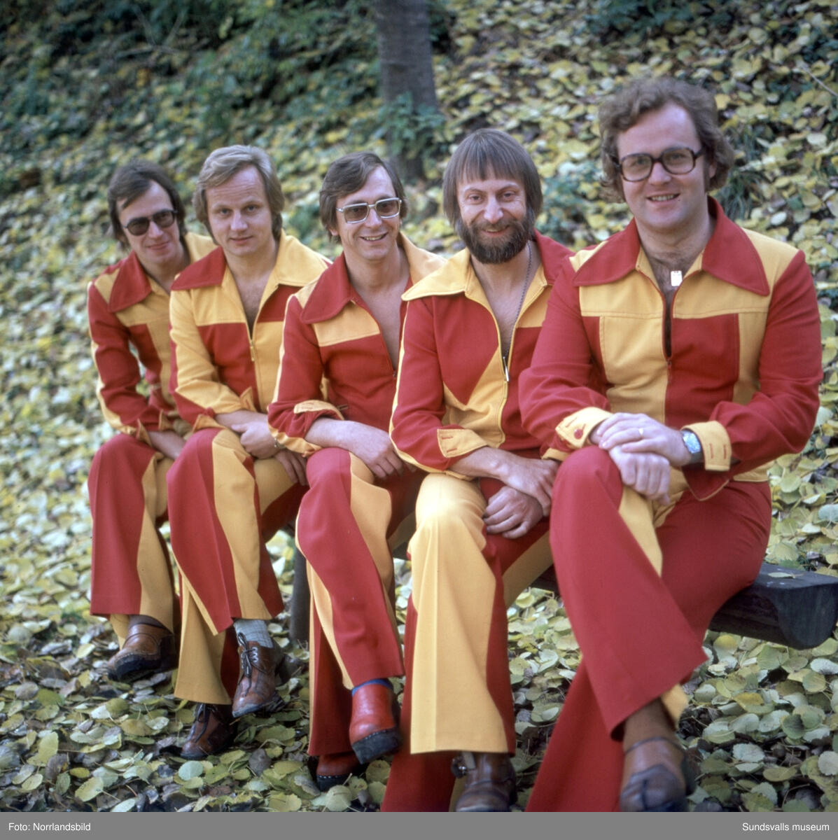 Fem personer sitter på en bänk i en park, klädda i matchande röda och gula kläder, omgivna av fallna gula löv.