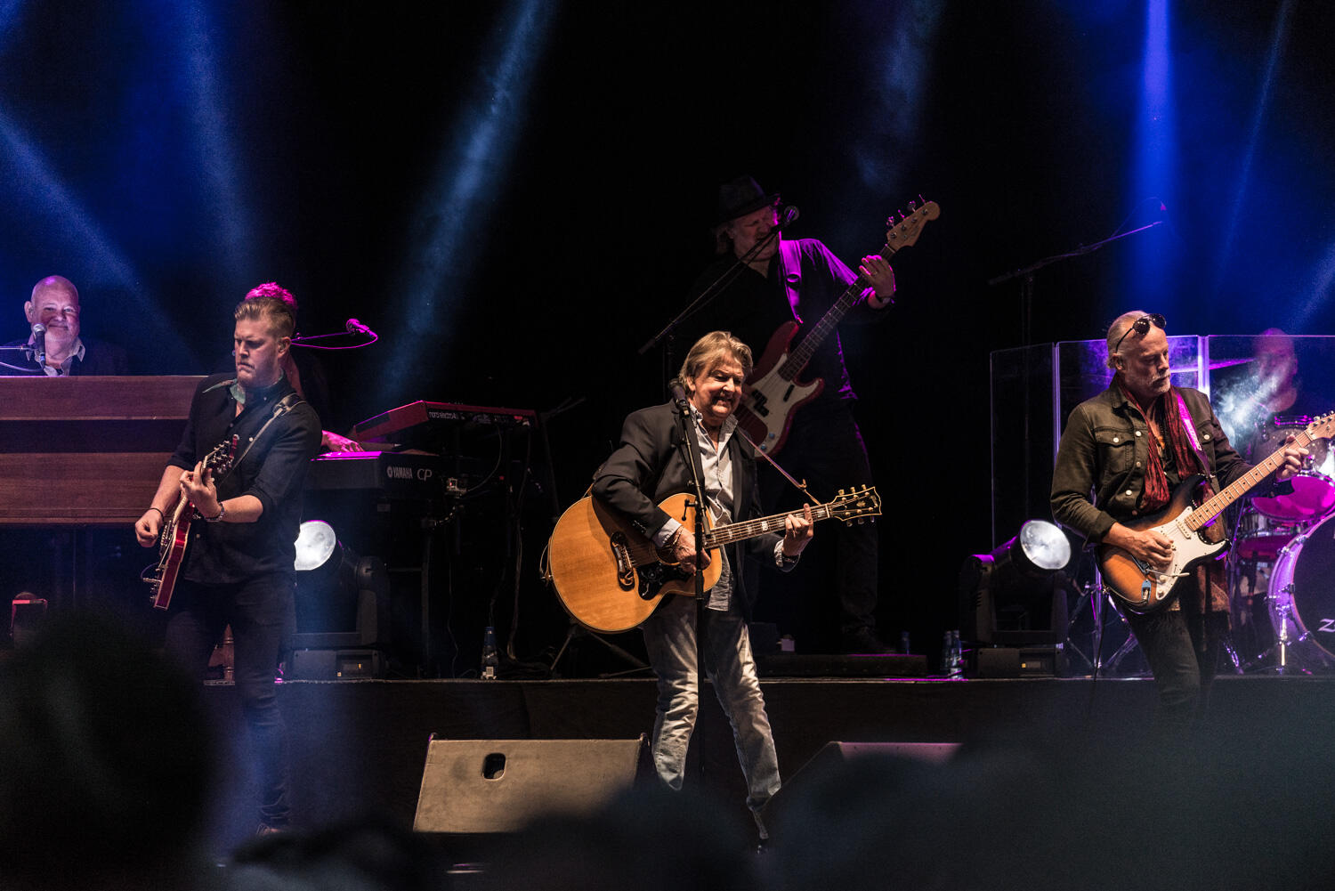 En grupp musiker uppträder på scenen med gitarrer, keyboard och livlig belysning, vilket skapar en energisk konsertatmosfär.