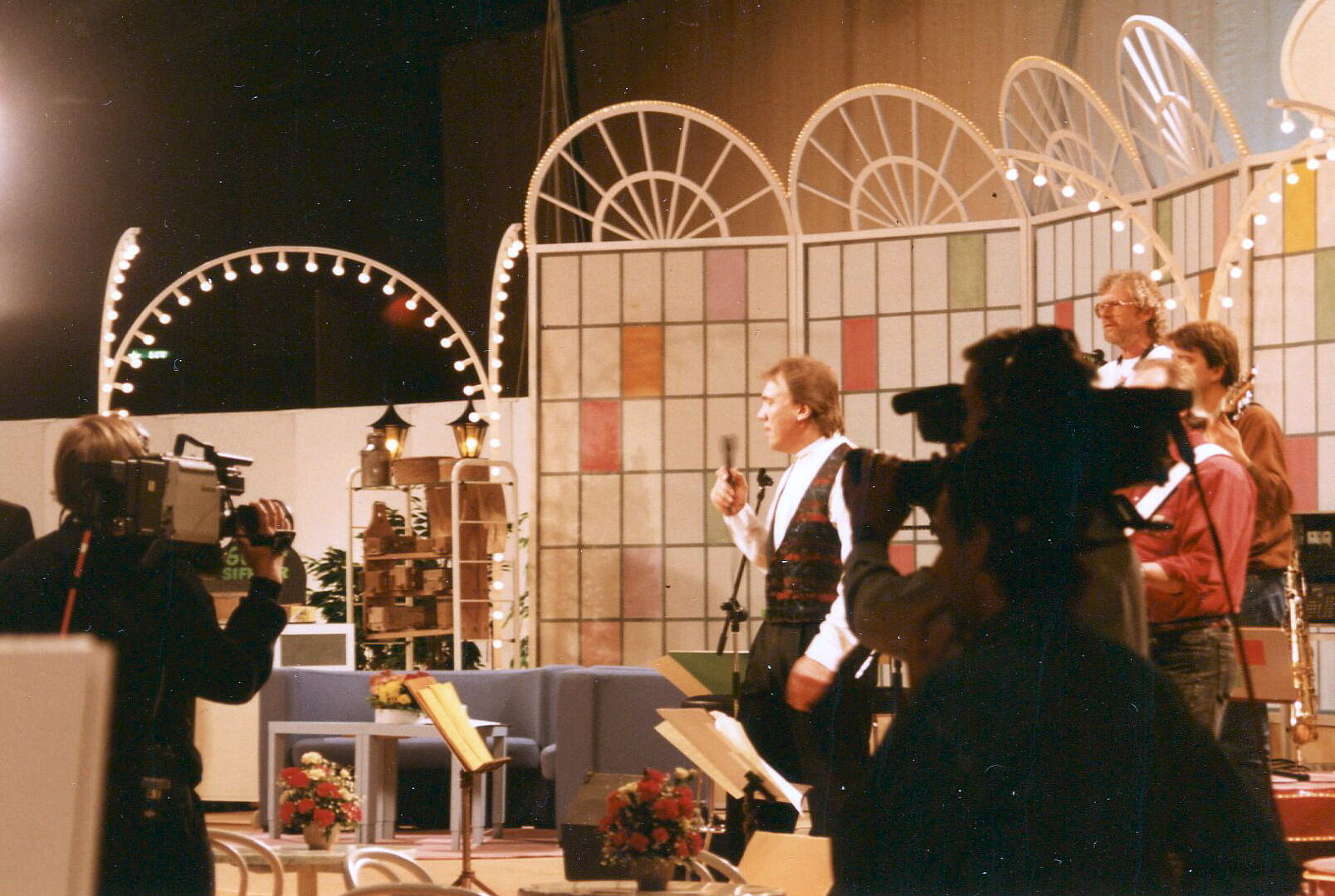 Tv-studio med artister på scenen, kameror som filmar, färgglad bakgrund och musikinstrument synliga.