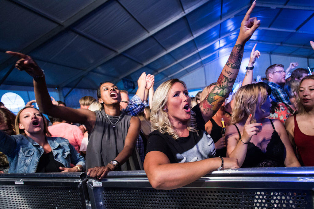 En livlig publik på en konsert, med händerna i luften, njuter av musiken under ett stort tält. Färgglada tatueringar och glada miner syns tydligt.