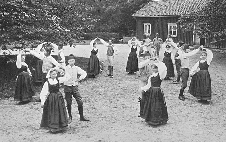 Svartvitt foto av människor i traditionella dräkter som dansar utomhus nära en rustik träbyggnad. Bild av Welin, Sanfrid via Digitalt museum. ID: 1M16-B145046:2741