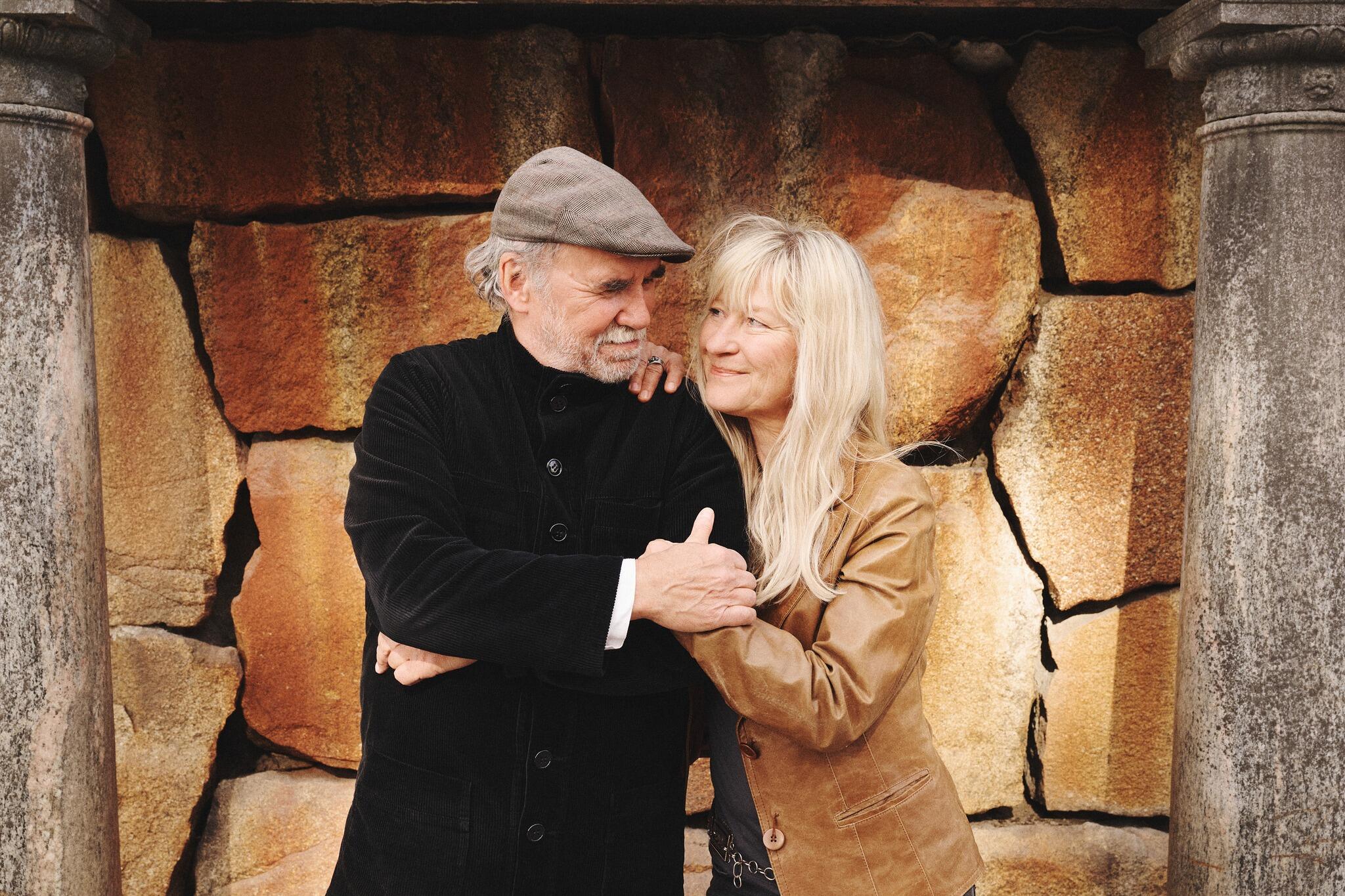 En man och en kvinna står tätt intill varandra mot en strukturerad stenvägg, ler och umgås varmt med varandra.