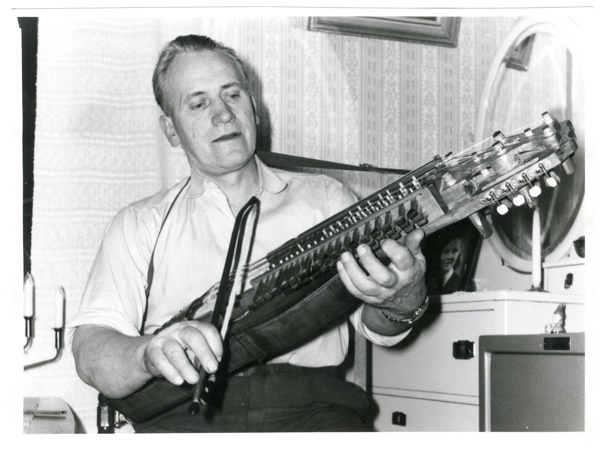 En person håller en nyckelharpa, sittande i ett rum med mönstrad tapet och en spegel i bakgrunden.