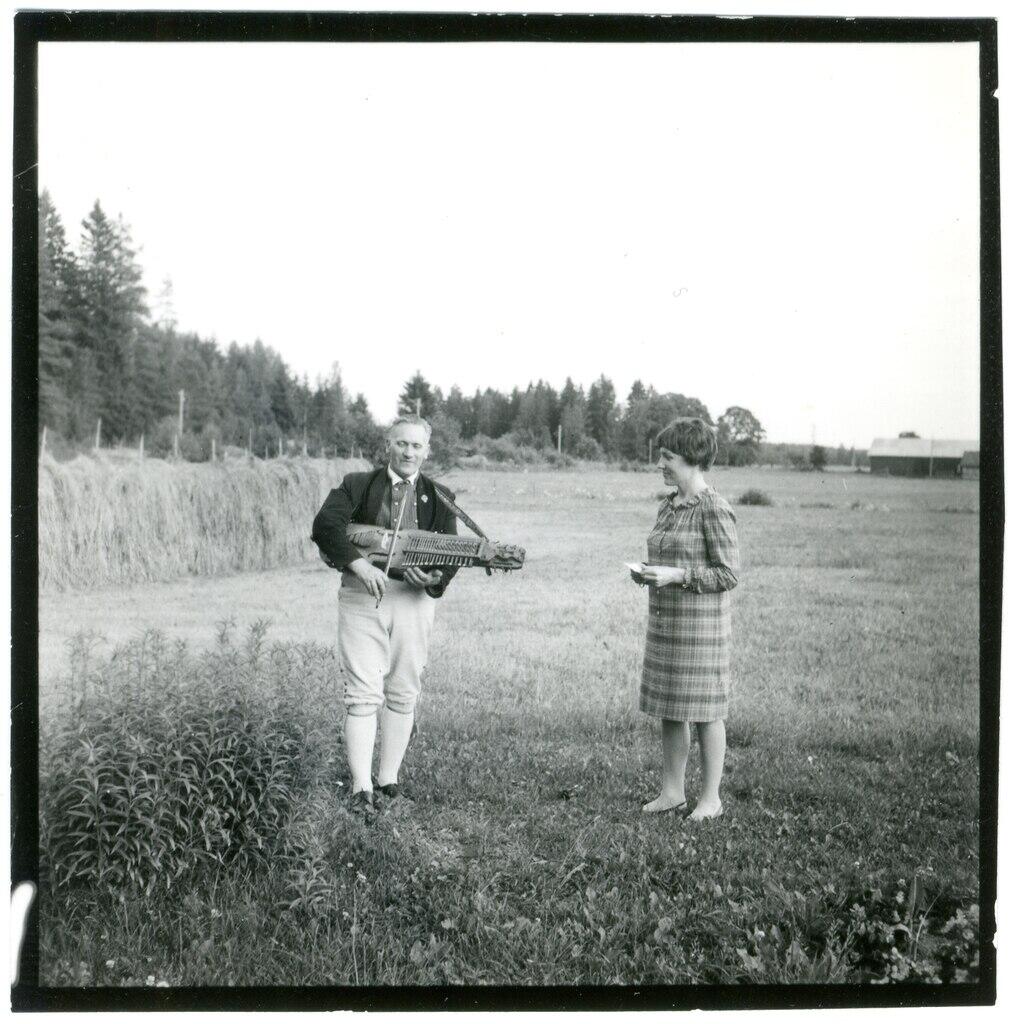 En person spelar ett instrument medan en annan står bredvid, båda på en gräsbevuxen äng omgiven av träd och en ladugård i bakgrunden.