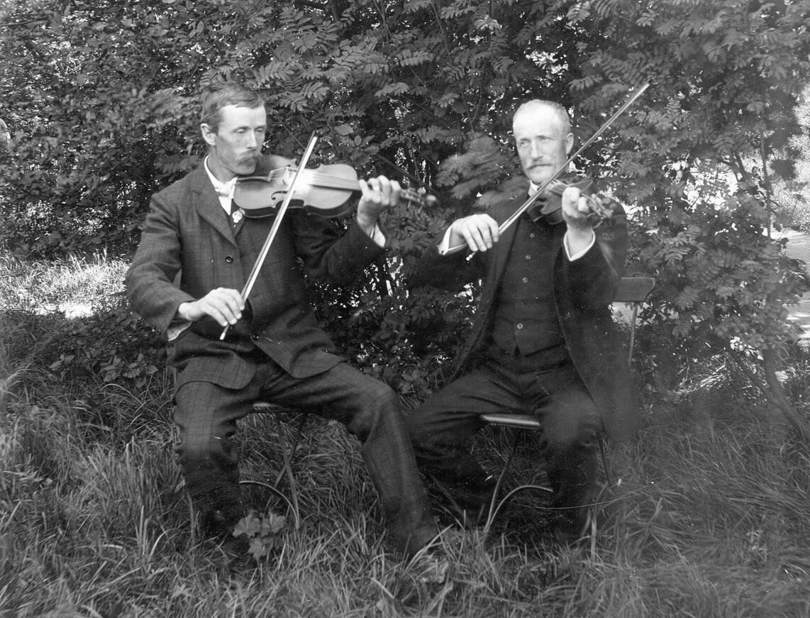 Två män spelar fiol medan de sitter på stolar utomhus, omgivna av frodig grönska. De bär formella kläder från en tidigare epok.