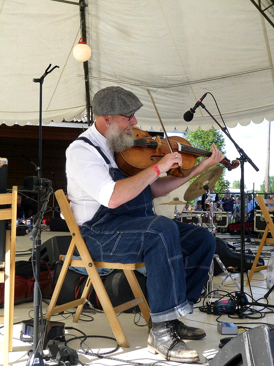 En musiker i overall spelar fiol under ett tält vid ett livligt utomhusevenemang, omgiven av musikutrustning och instrument.