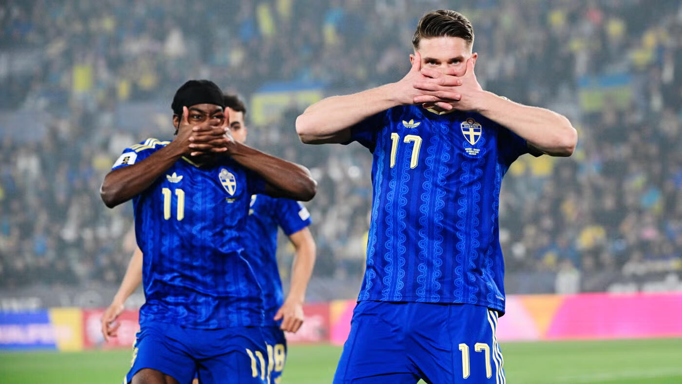 Två svenska fotbollsspelare i blå landslagströjor firar ett mål. Spelaren med tröjnummer 17 gör en målgest där han håller händerna för munnen. I bakgrunden syns en fullsatt publikläktare i gult och blått.
