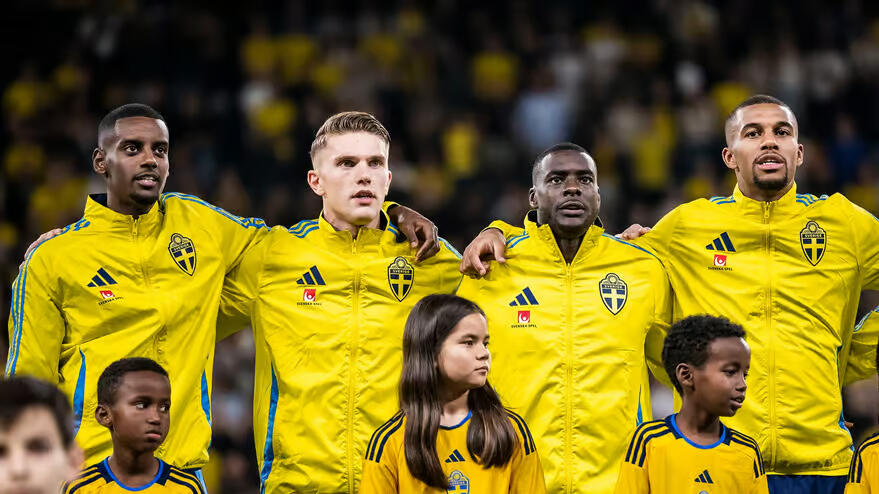 Fyra svenska fotbollsspelare i gula landslagsjackor står uppradade och sjunger nationalsången. I förgrunden syns tre barn i gula tröjor. Publiken i bakgrunden är iklädd gult.