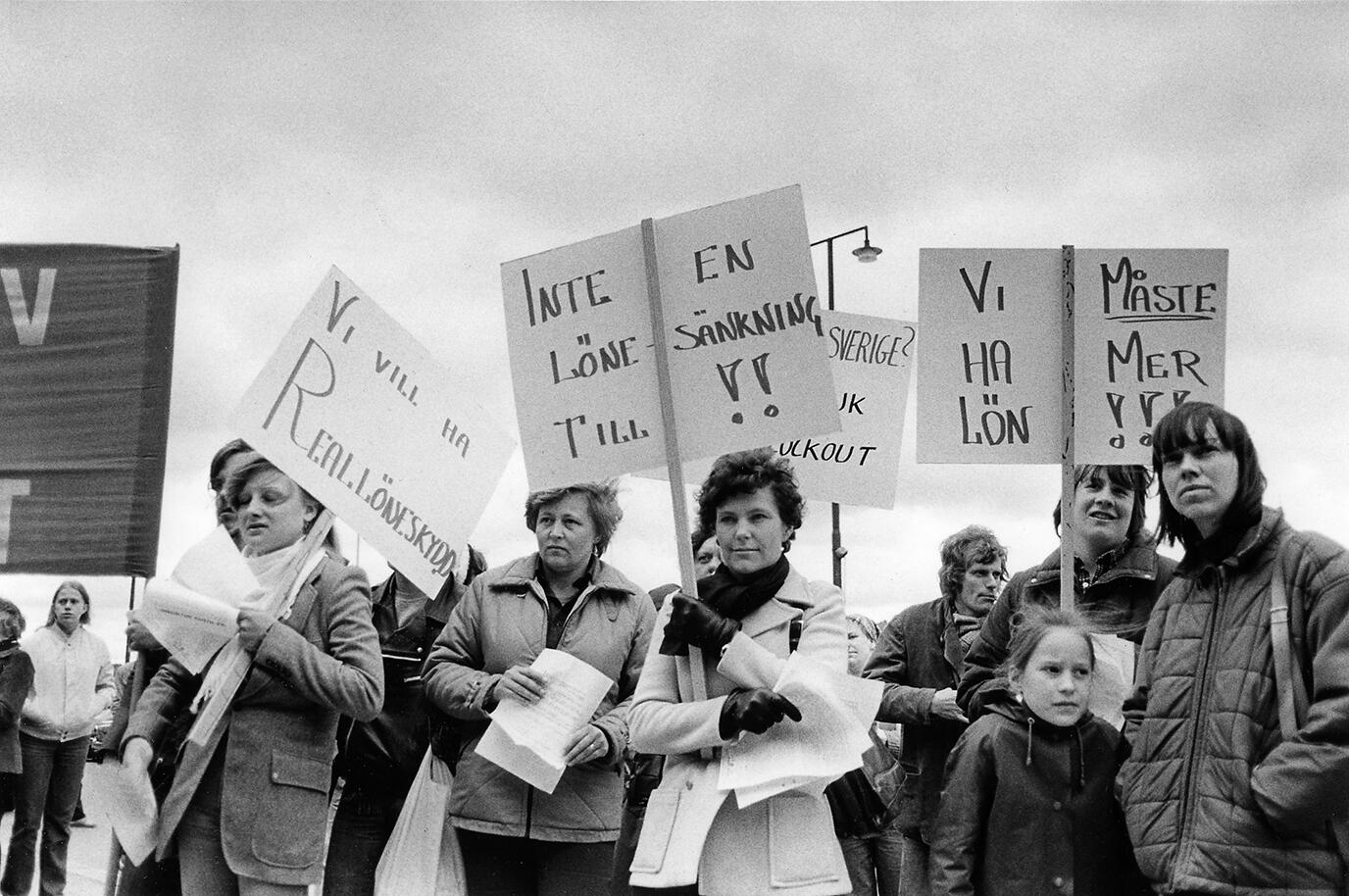 Svartvit bild från en löneprotest med huvudsakligen kvinnor som håller handskrivna plakat med krav som 