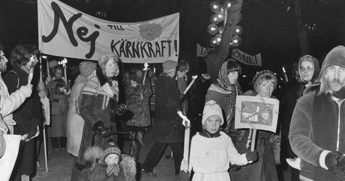 Svartvit bild från fackeltåg mot kärnkraft, med stor banderoll där det står "Nej till kärnkraft" med en leende sol. Demonstranter i vinterkläder, bland annat barn, går med tända facklor.