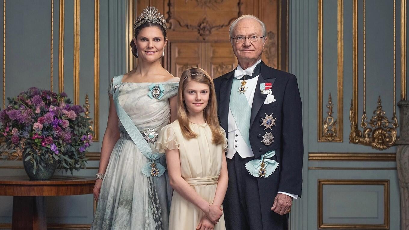 Formellt gruppfoto av tre medlemmar ur svenska kungafamiljen i en praktfull salong. En kvinna i ljus aftonklänning med tiara och ordensband, en ung flicka i ljusgul klänning, och en äldre man i mörk kavaj med ordnar och band. Blomsterarrangemang och guldornamenterade väggpaneler i bakgrunden.