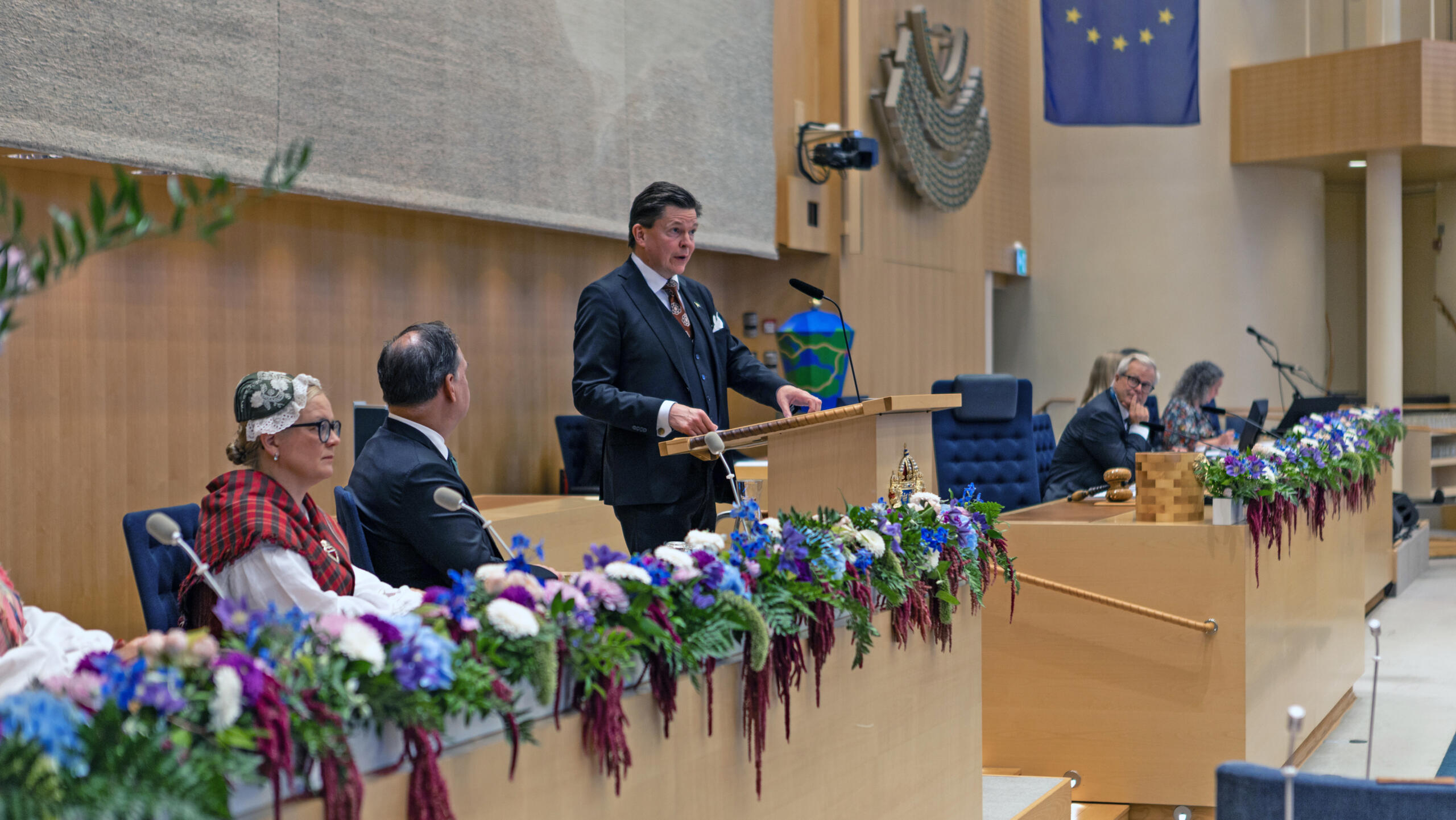 Talmannen håller tal från talarstolen i Sveriges riksdags plenisal vid riksmötets öppnande, med sittande riksdagsledamöter i förgrunden. Talarstolen är dekorerad med blomsterarrangemang i blått och lila, och en EU-flagga syns i bakgrunden. En kvinna i traditionell dräkt sitter bland åhörarna.
