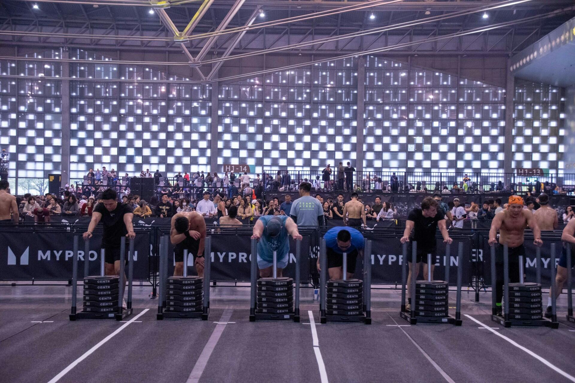 Deltagare på en HYROX-tävling står på rad bakom tunga, viktlastade slädar och förbereder sig för sled push, medan publik tittar på bakom avspärrningarna.