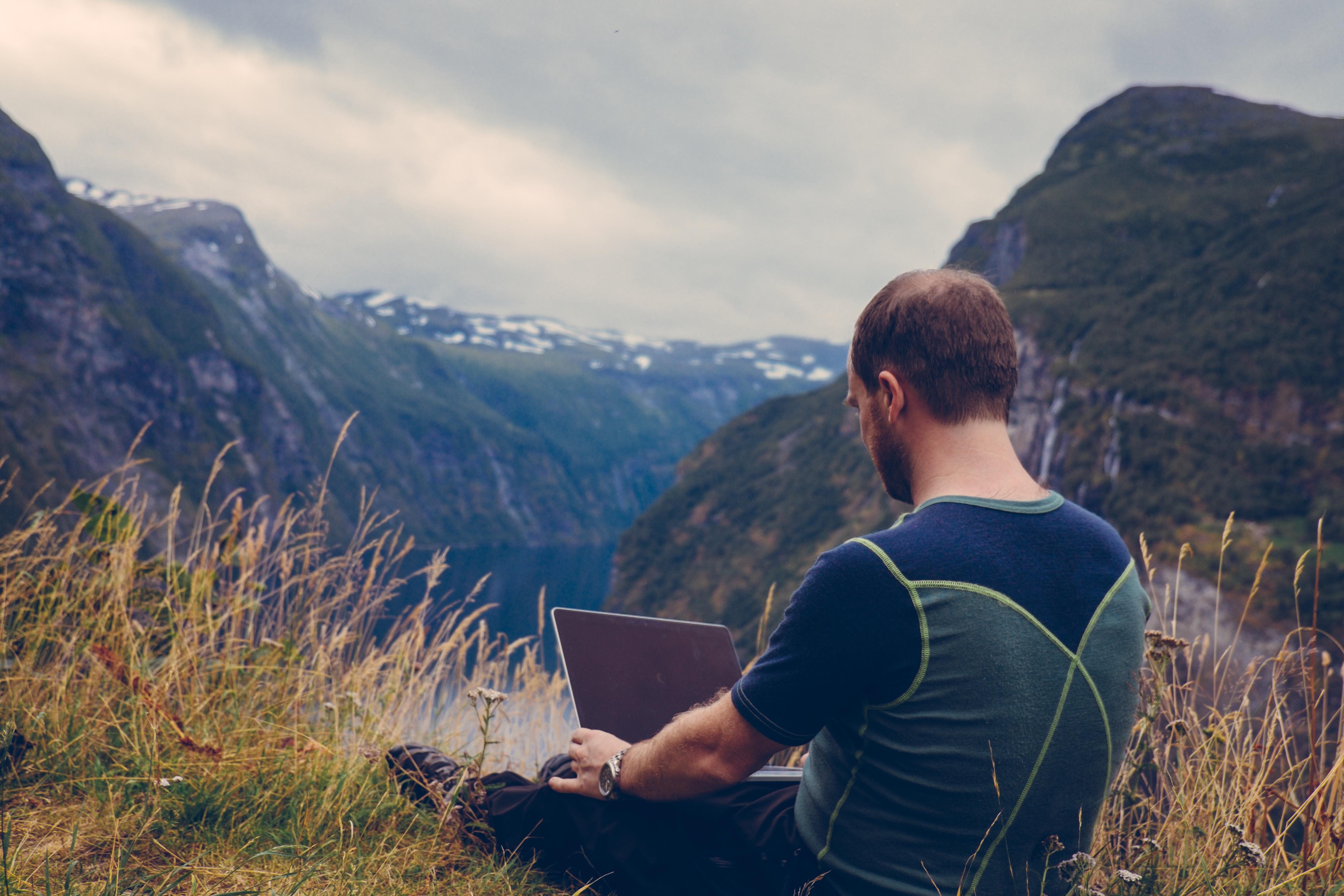 A person in a green and blue short-sleeved shirt sits atop a mountain with an open laptop computer on his lap, facing a valley and other peaks in the distance.