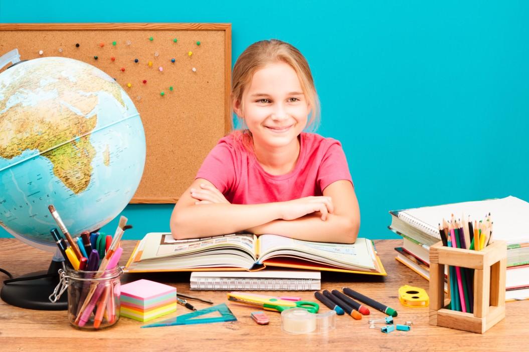 A girl in a pink top smiles as sits at a desk laden with learning tools such as books, writing instruments and a globe.