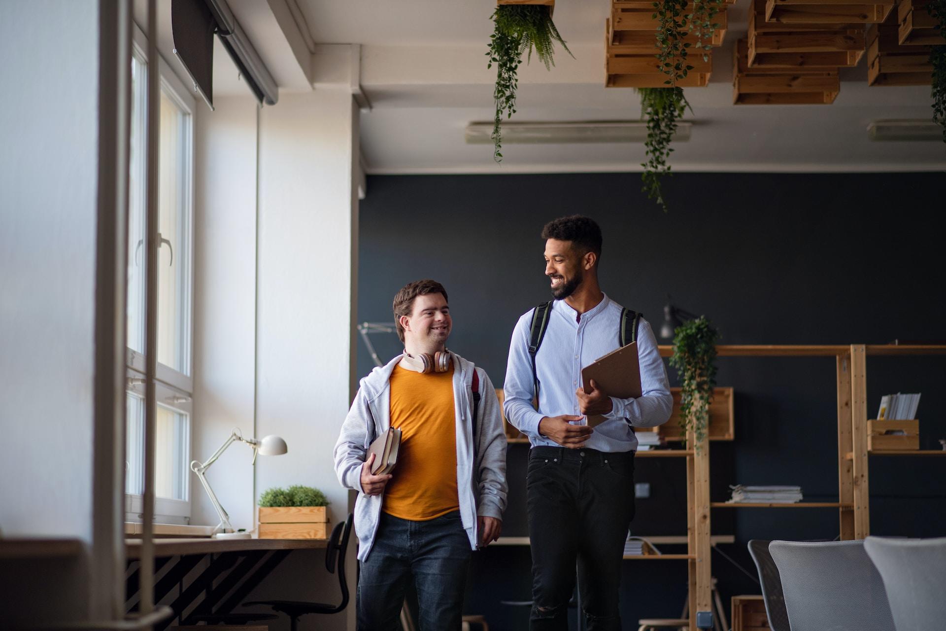 A person wearing a light blue button-down shirt and carrying a backpack holds books in their arm while walking alongside a person in an orange shirt who is also carrying books. 
