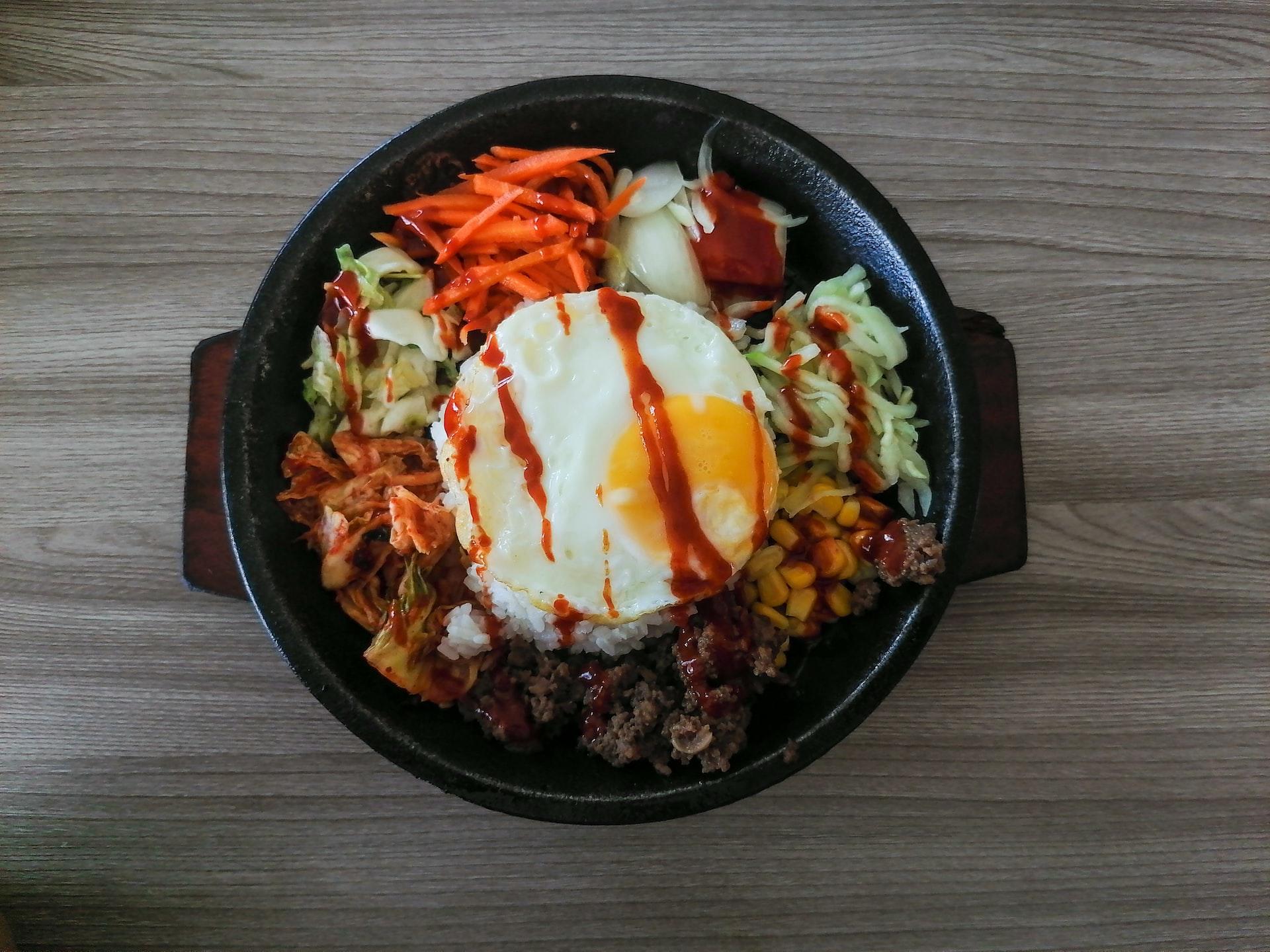 a black bowl containing variously-coloured vegetables surrounding a fried egg that is drizzled in chili paste rests on a wooden surface. 