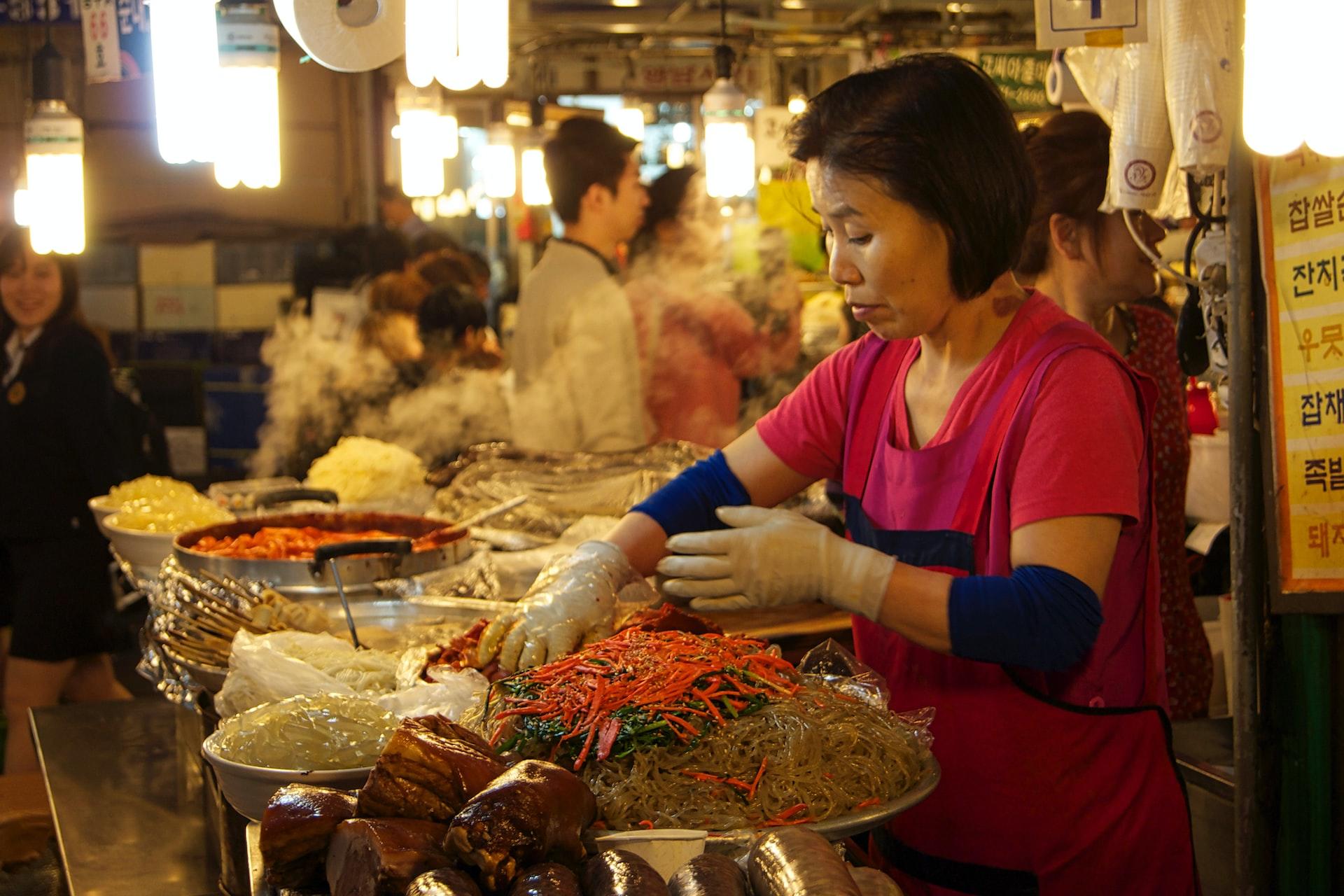 A woman in a short-sleeved orange top, wearing food handler gloves, tends to her food bar in a lit diner while patrons look on.