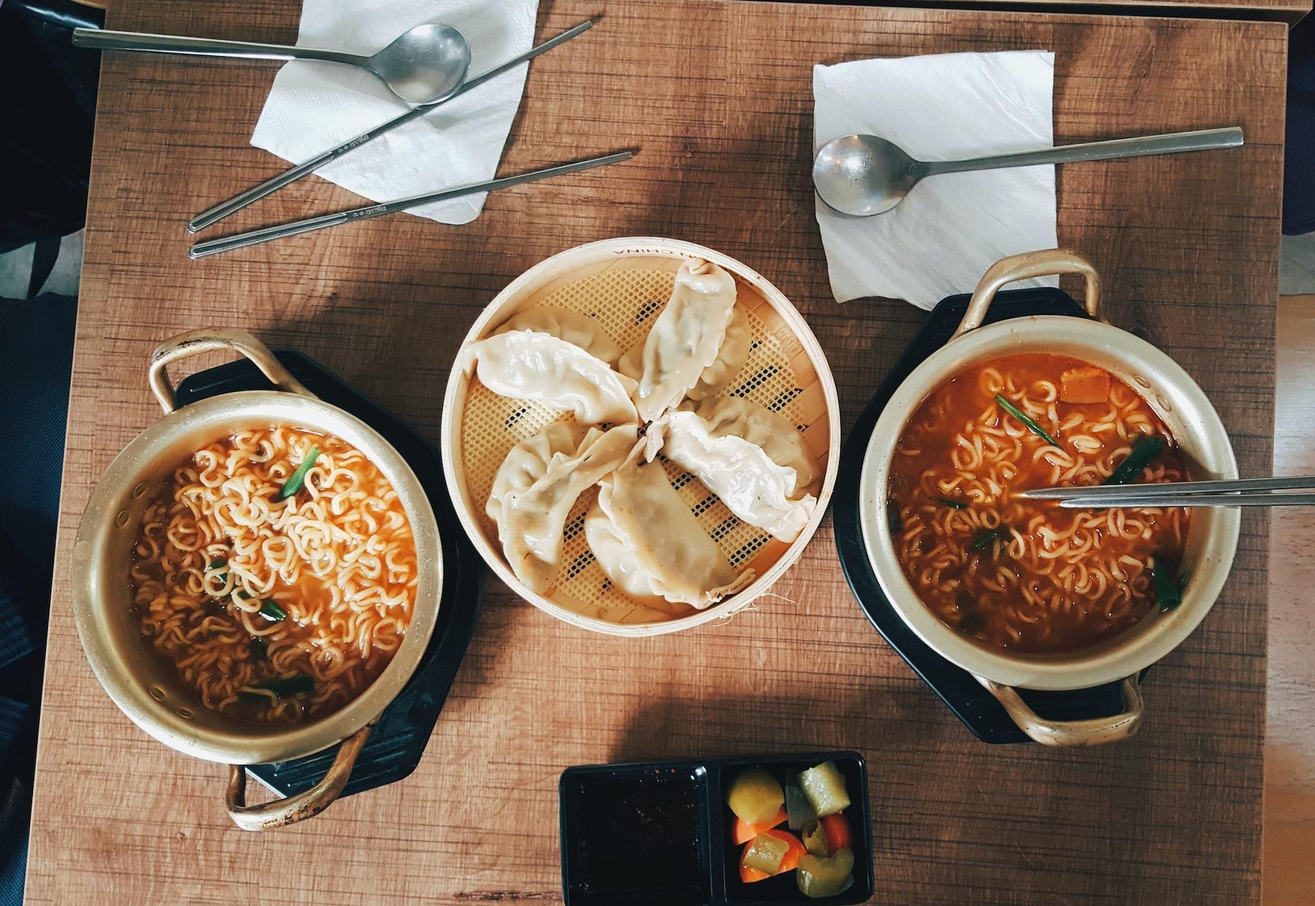 A Korean table set with two bowls of ramyeon and a steamer basked of dumplings with spoons and chopsticks at the ready. 