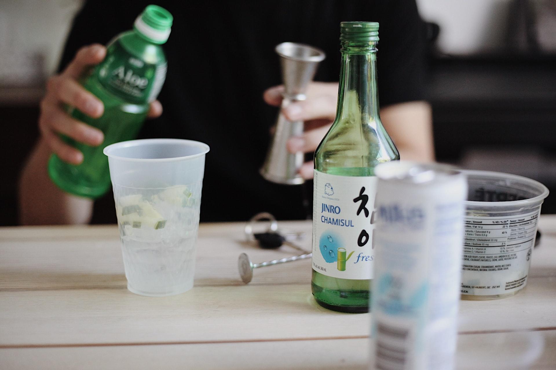 A person in a black top holding a green bottle in one hand and a jigger in the other behind a table upon which are a plastic cup, a bottle of soju and a can of mixer.