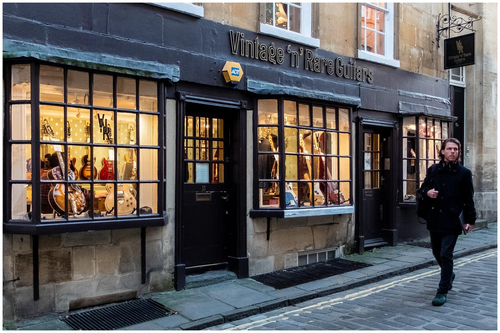 A guitar shop with a name board Vintage 'n' Rare Guitars showcasing their guitar collections by the widows of the shop. A man in black clothes is seen passing by holding his laptop. 