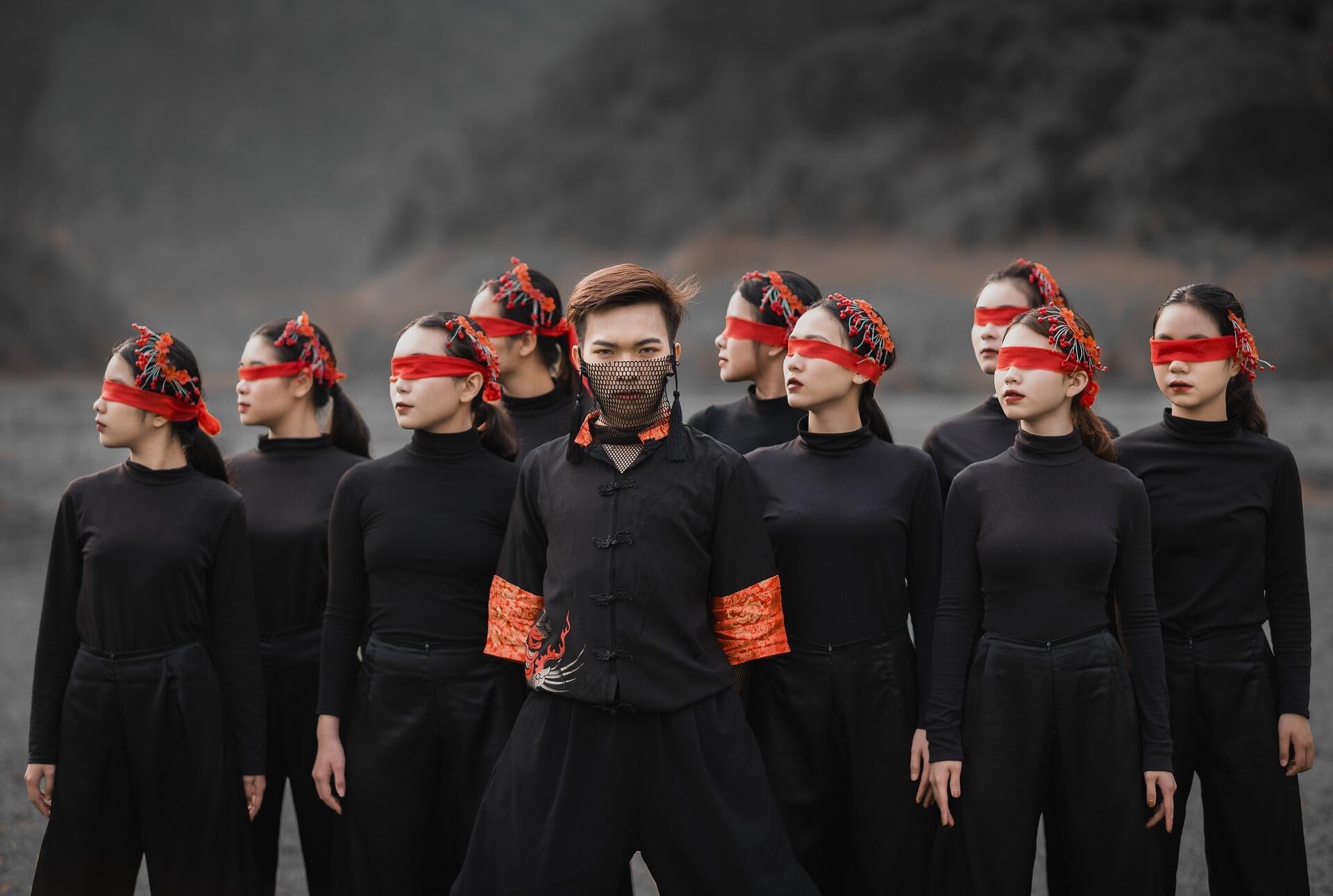 Dancers in black outfits with red headpieces pose boldly against a dramatic backdrop.