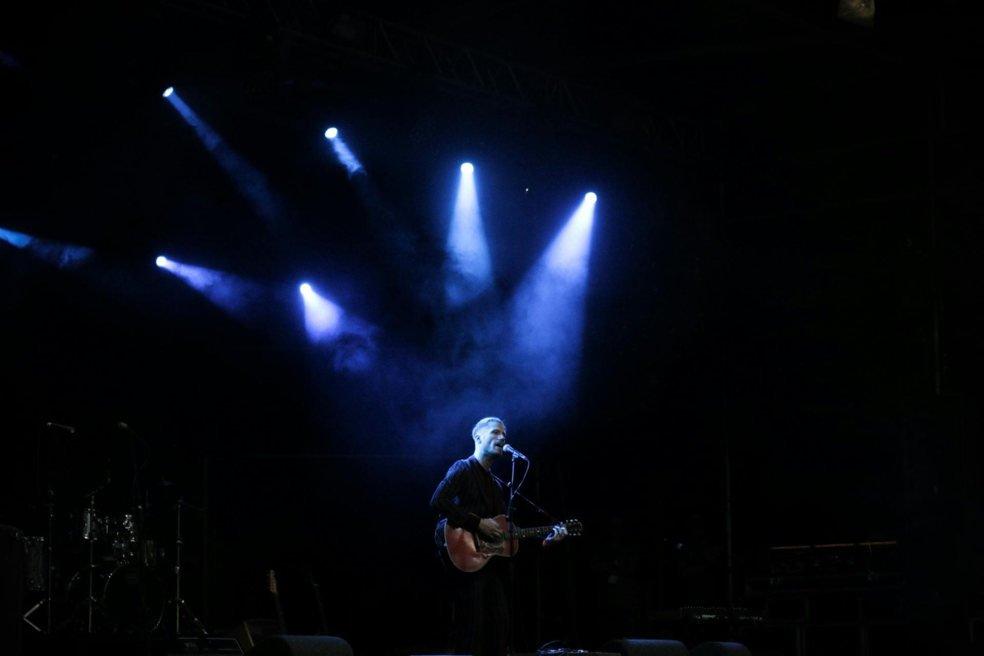  A man is seen performing on stage singing while strumming his guitar. 