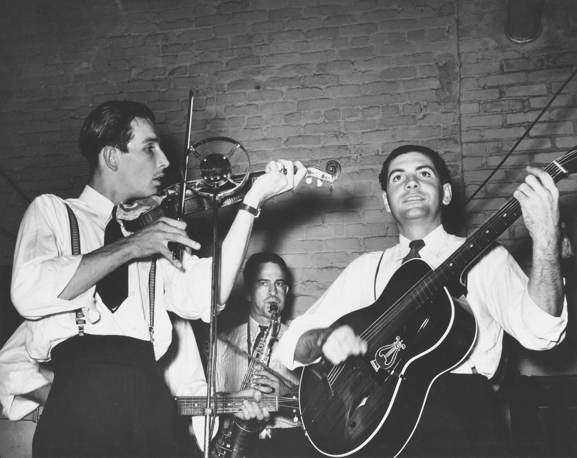 Three musicians in formal attire performing with violin, saxophone, and acoustic guitar in black and white vintage photograph