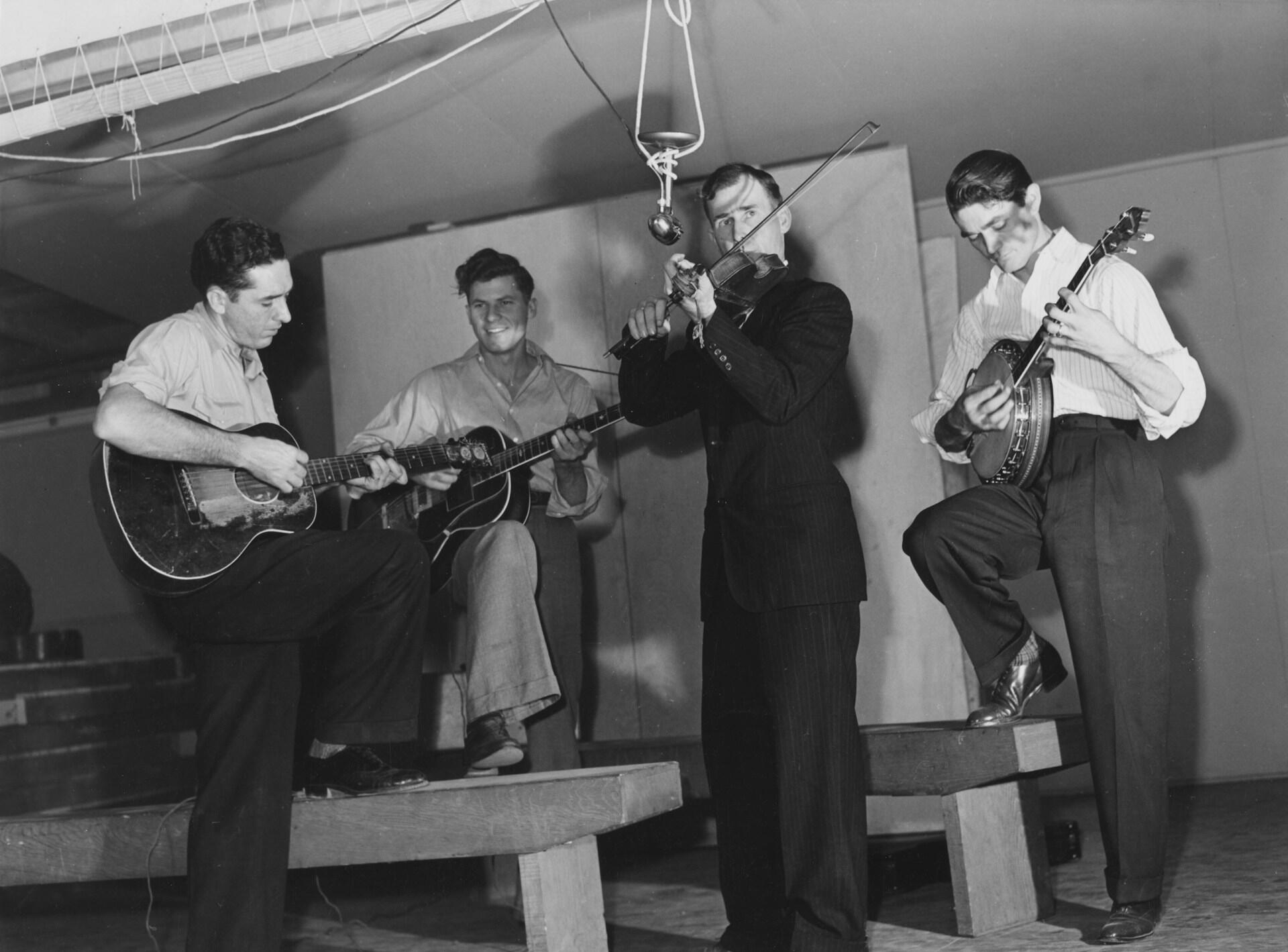 Four musicians performing in vintage black and white photograph with acoustic guitars, violin, and banjo in what appears to be a recording studio or performance space