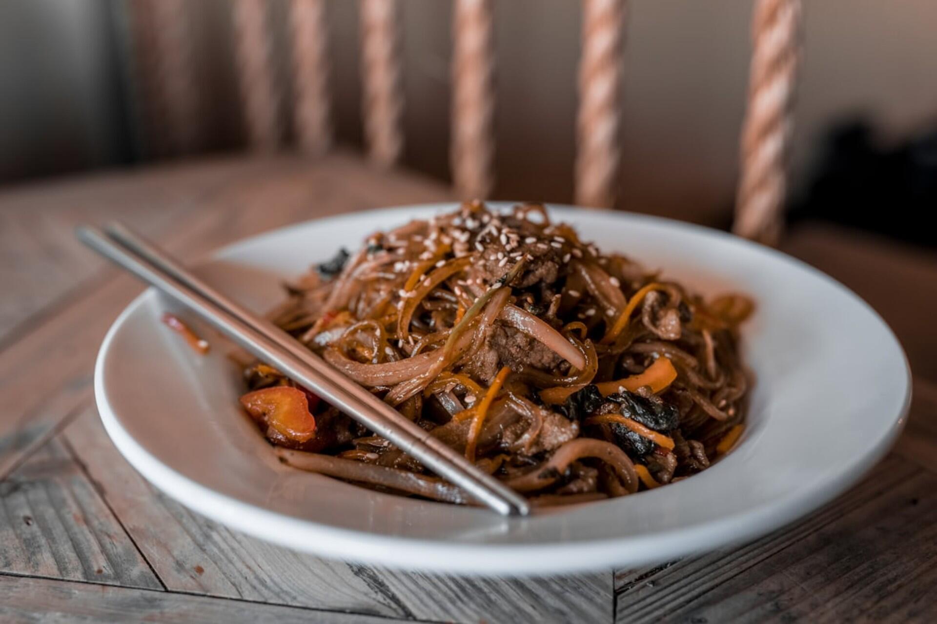 Close-up of japchae, Korean glass noodles stir-fried with beef, carrots, onions, and sesame seeds, served on a white plate with chopsticks.