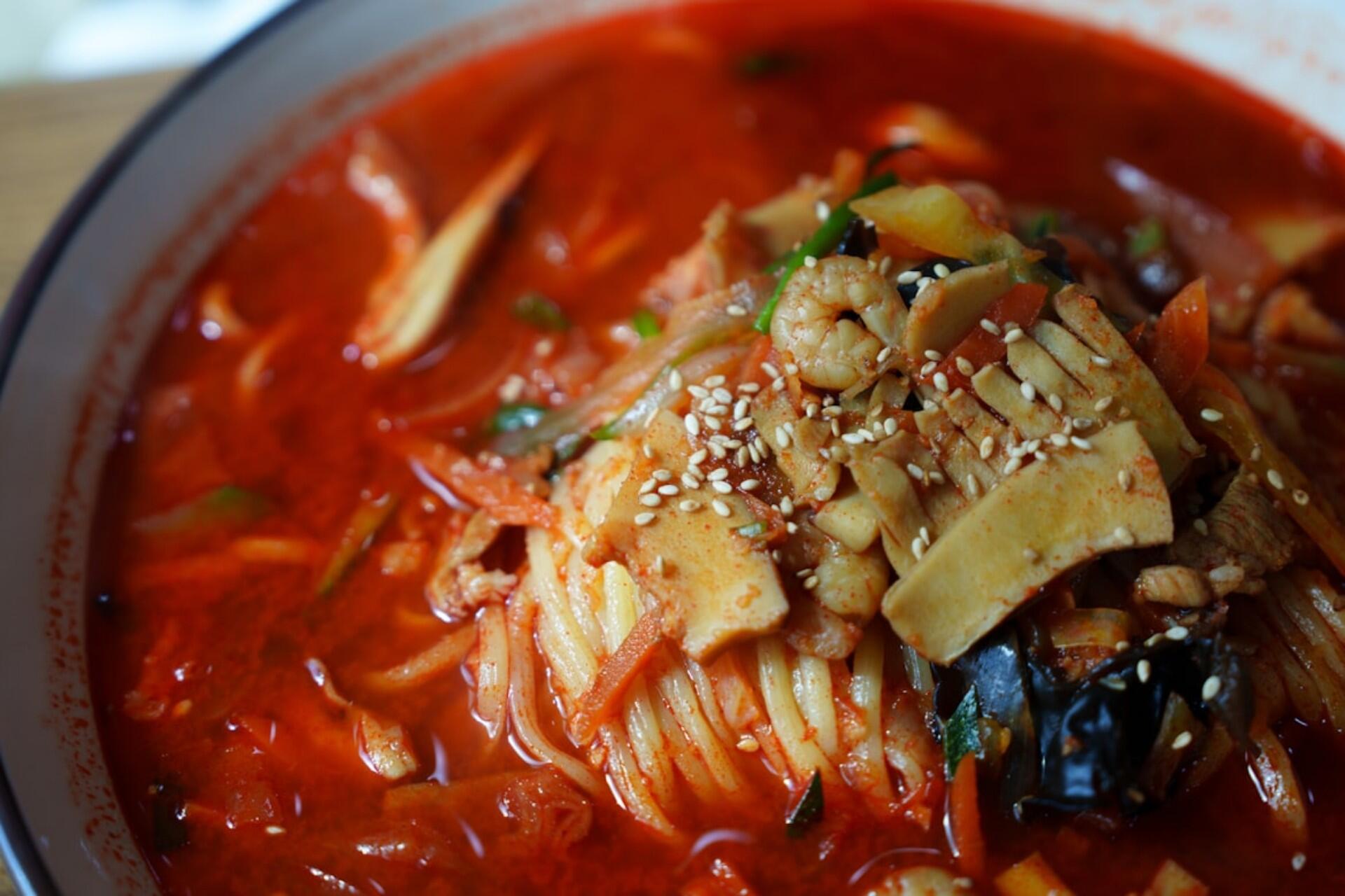 Close-up of kimchi stew with noodles, sliced vegetables, seafood, and sesame seeds in a rich red broth.