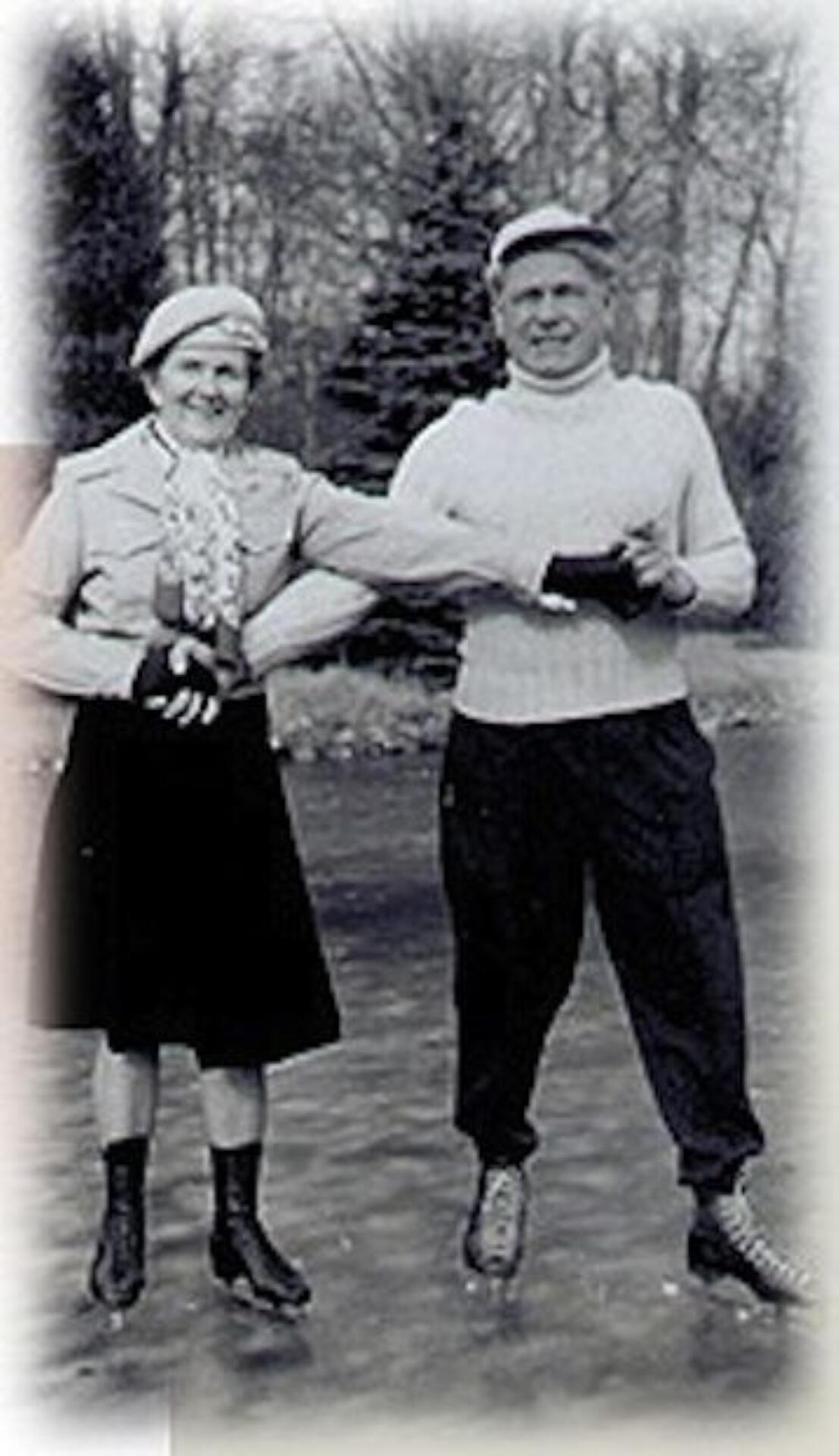Two individuals stand on a frozen lake, dressed warmly, with snow-covered trees in the background.