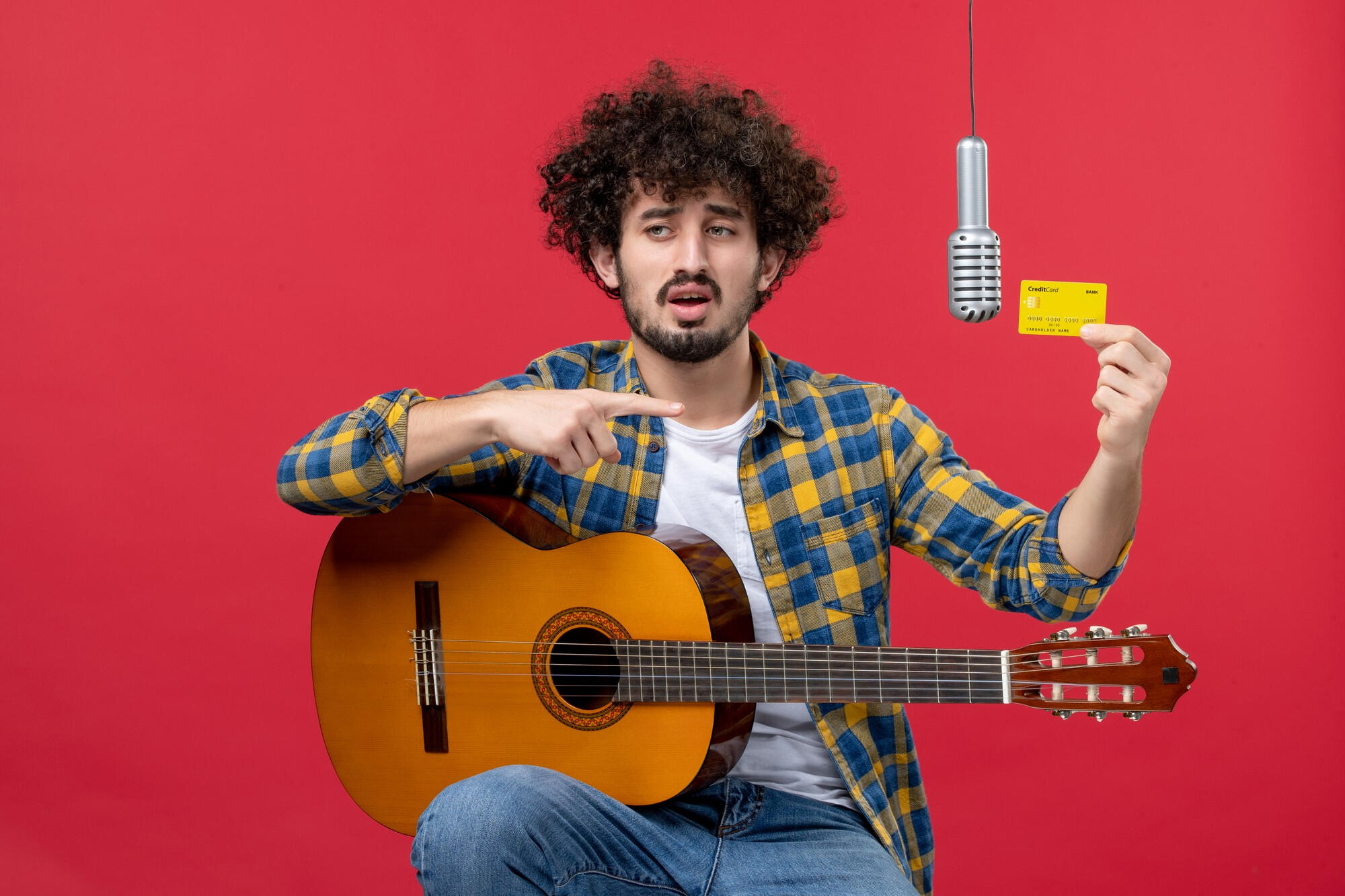 Young man with curly hair holding a credit card and pointing at it while sitting with an acoustic guitar and a microphone on a red background.