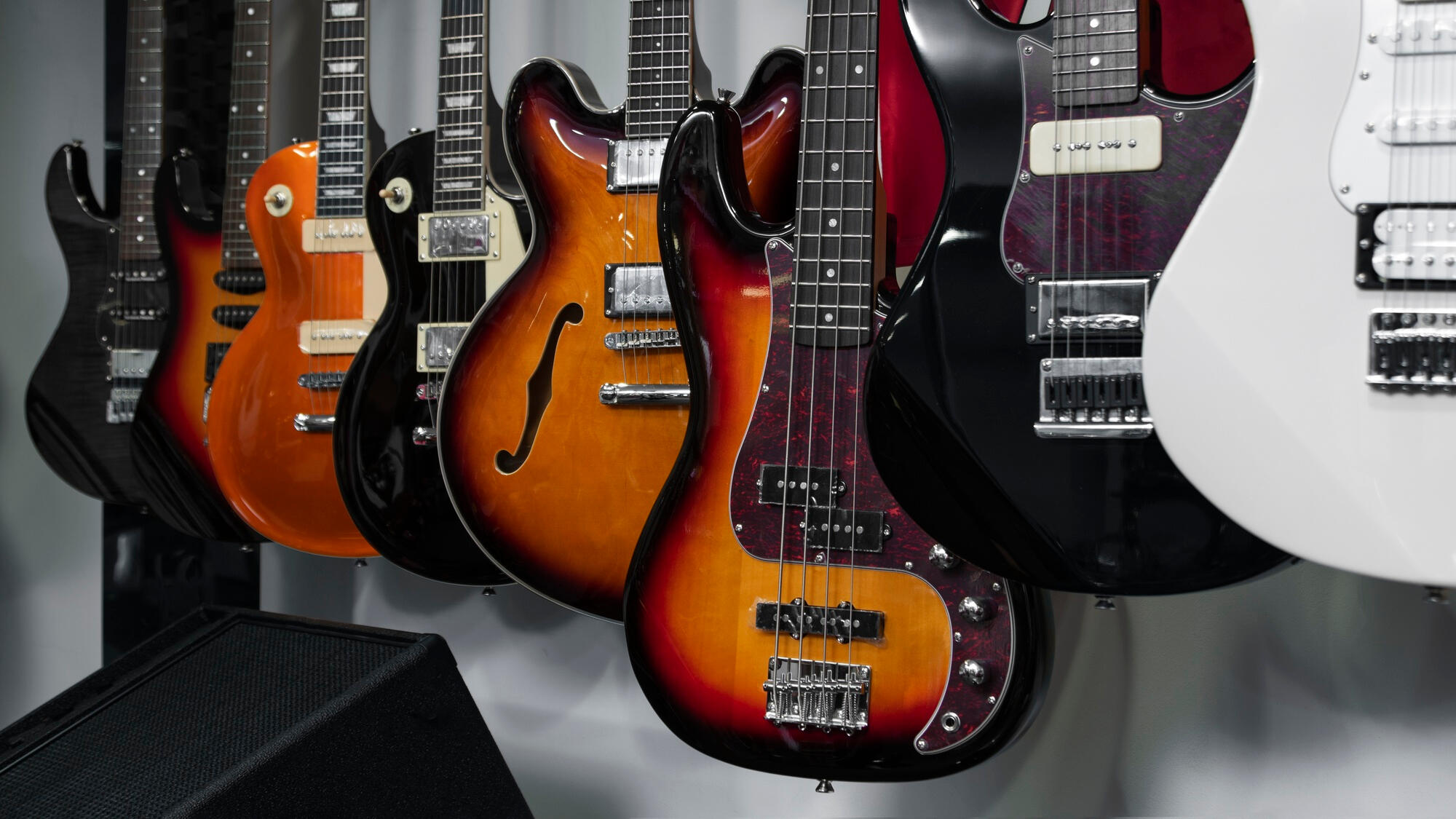 Row of various electric guitars and a bass guitar hanging on a wall above a black amplifier.