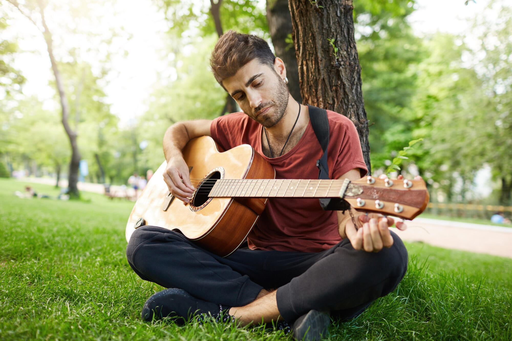 Young man sitting cross-legged under a tree in a park, playing an acoustic guitar on a sunny day.