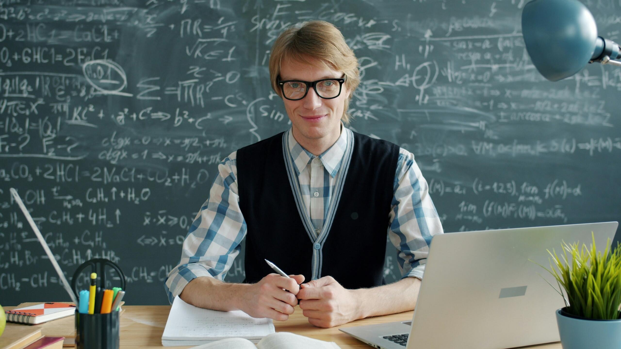 A male teacher smiling at his desk in front of a blackboard.