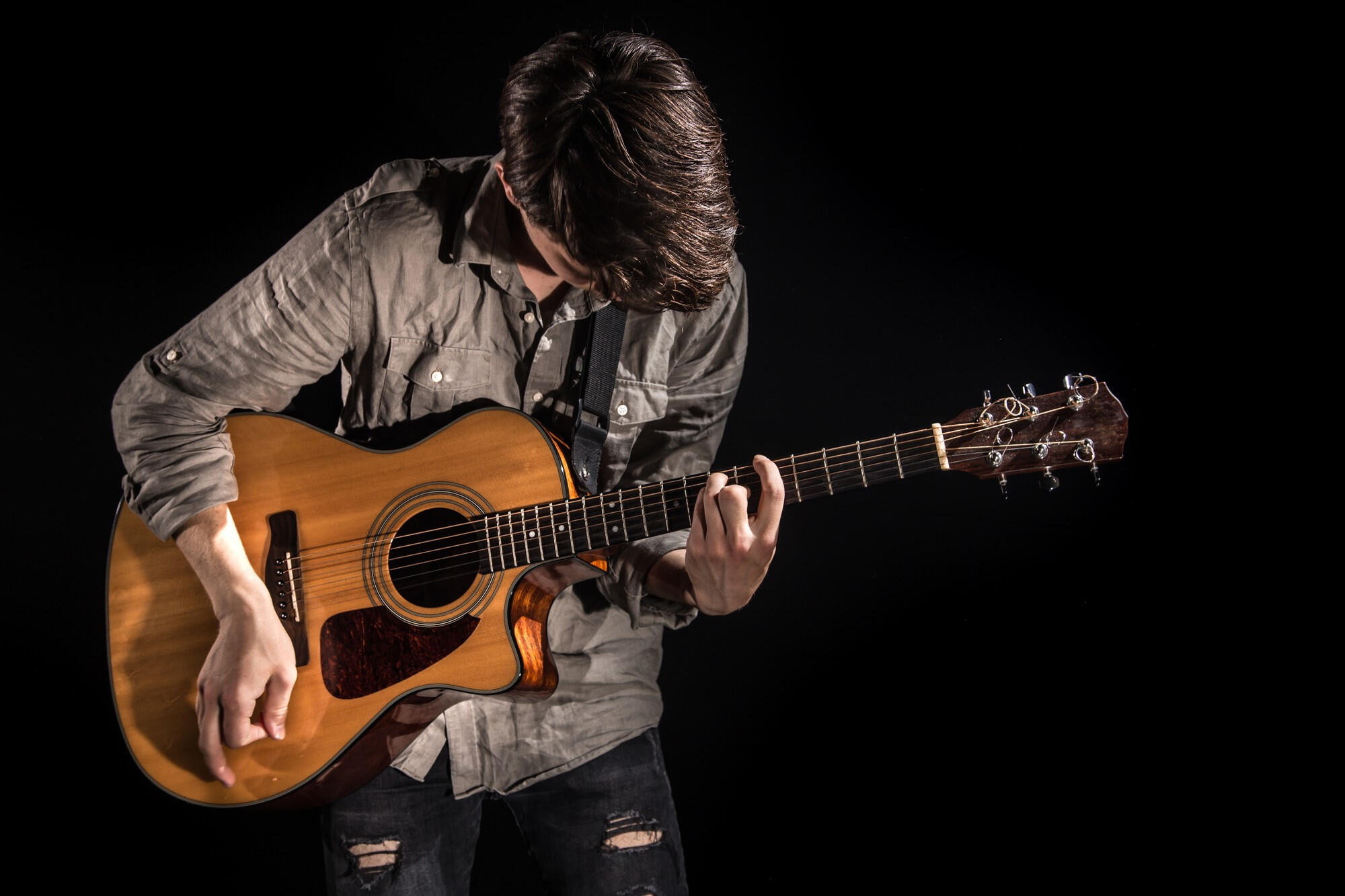 A young man wearing a gray shirt plays an acoustic guitar against a black background.