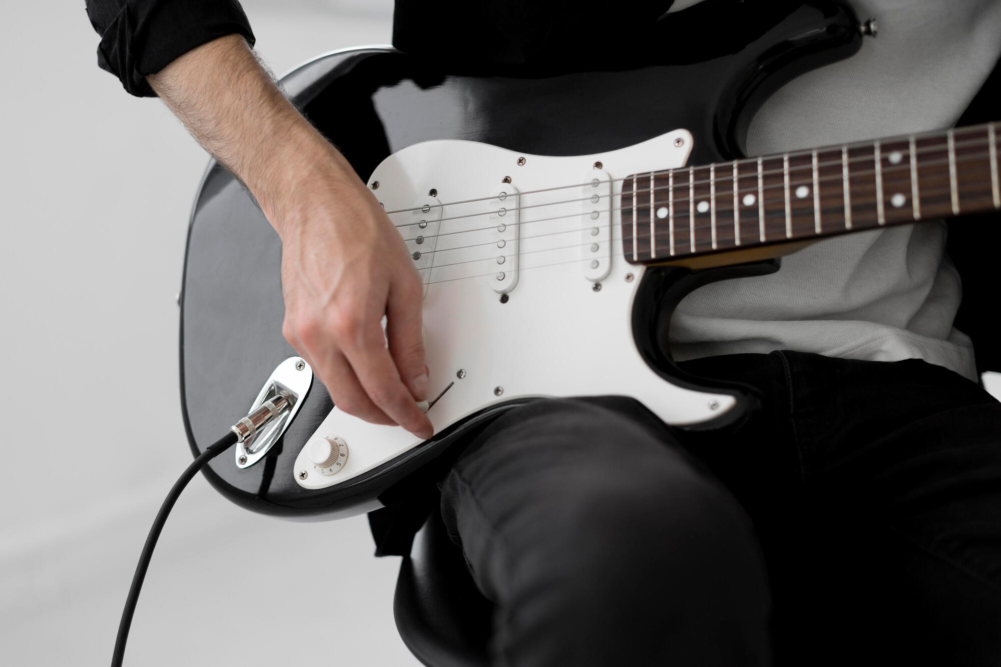 Close-up of a person playing a black and white electric guitar while seated, focusing on the hand strumming the strings.