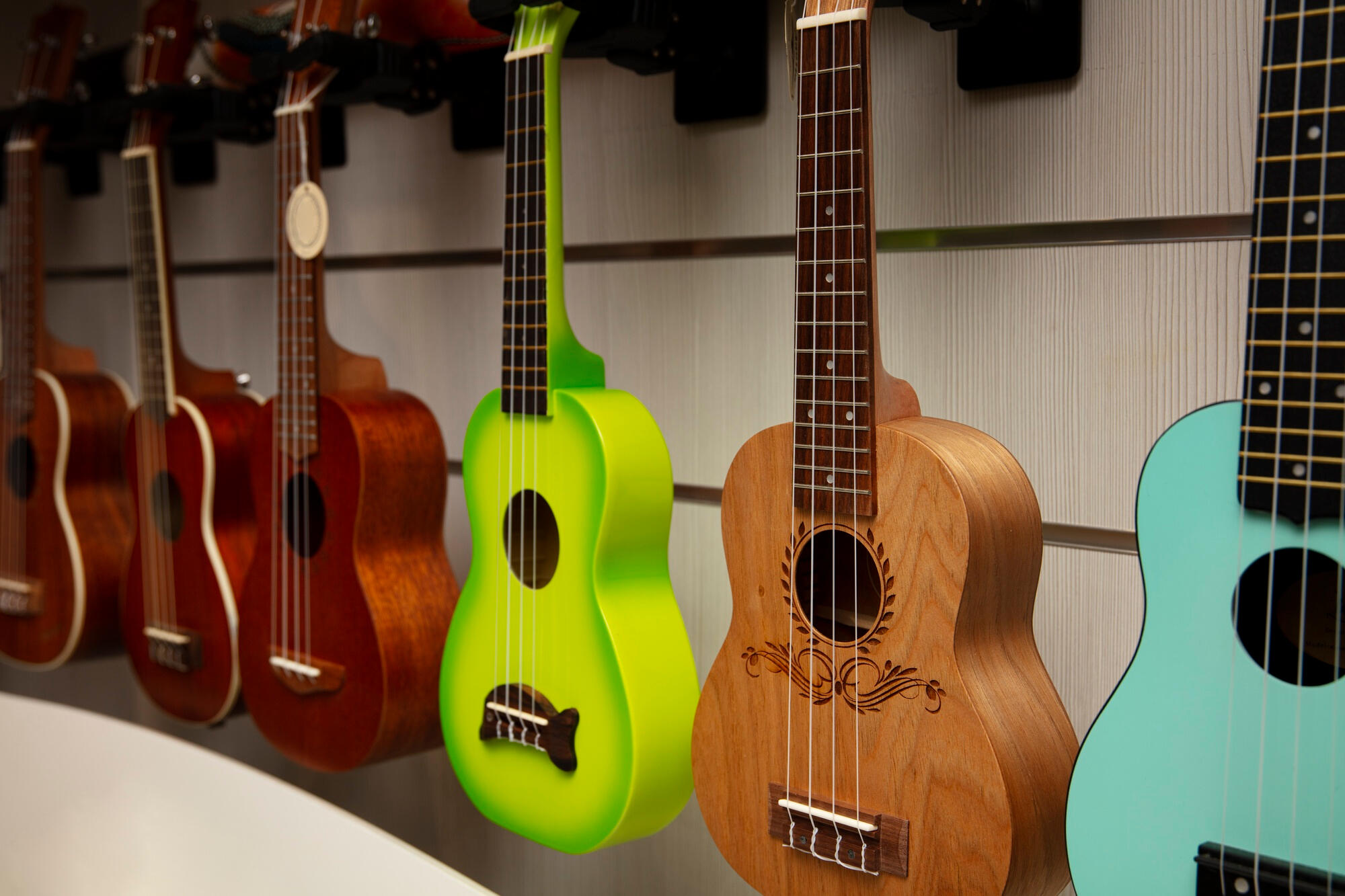 Row of colorful ukuleles hanging on a wall, featuring wooden, lime green, and blue finishes with various designs.