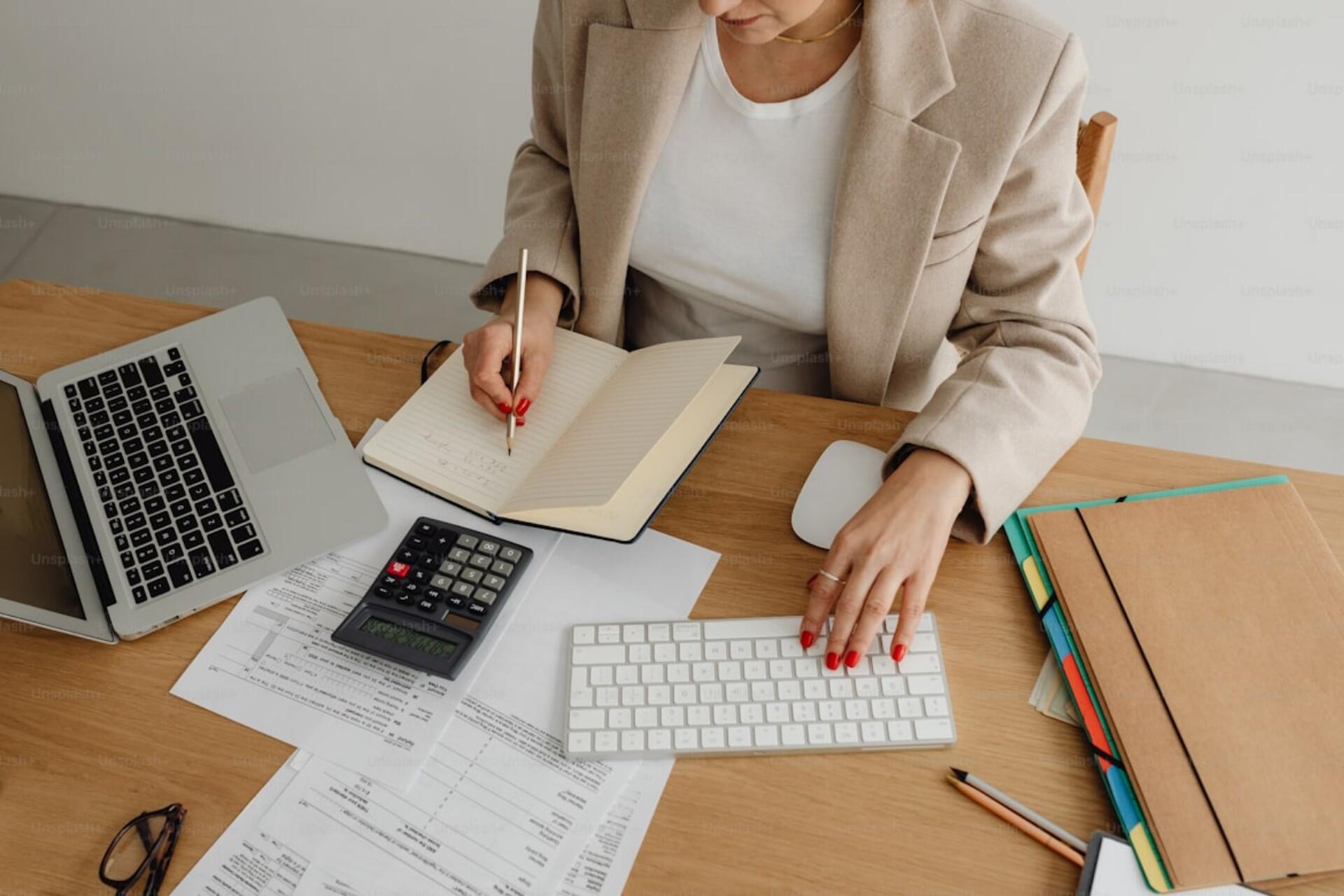Close-up of a woman in a beige blazer writing in a notebook while using a computer keyboard, with financial documents, a calculator, and folders on the desk