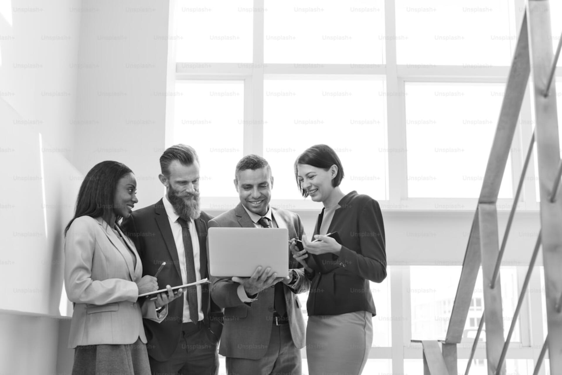 Group of business professionals in formal attire standing together and reviewing information on a laptop in a bright office space.