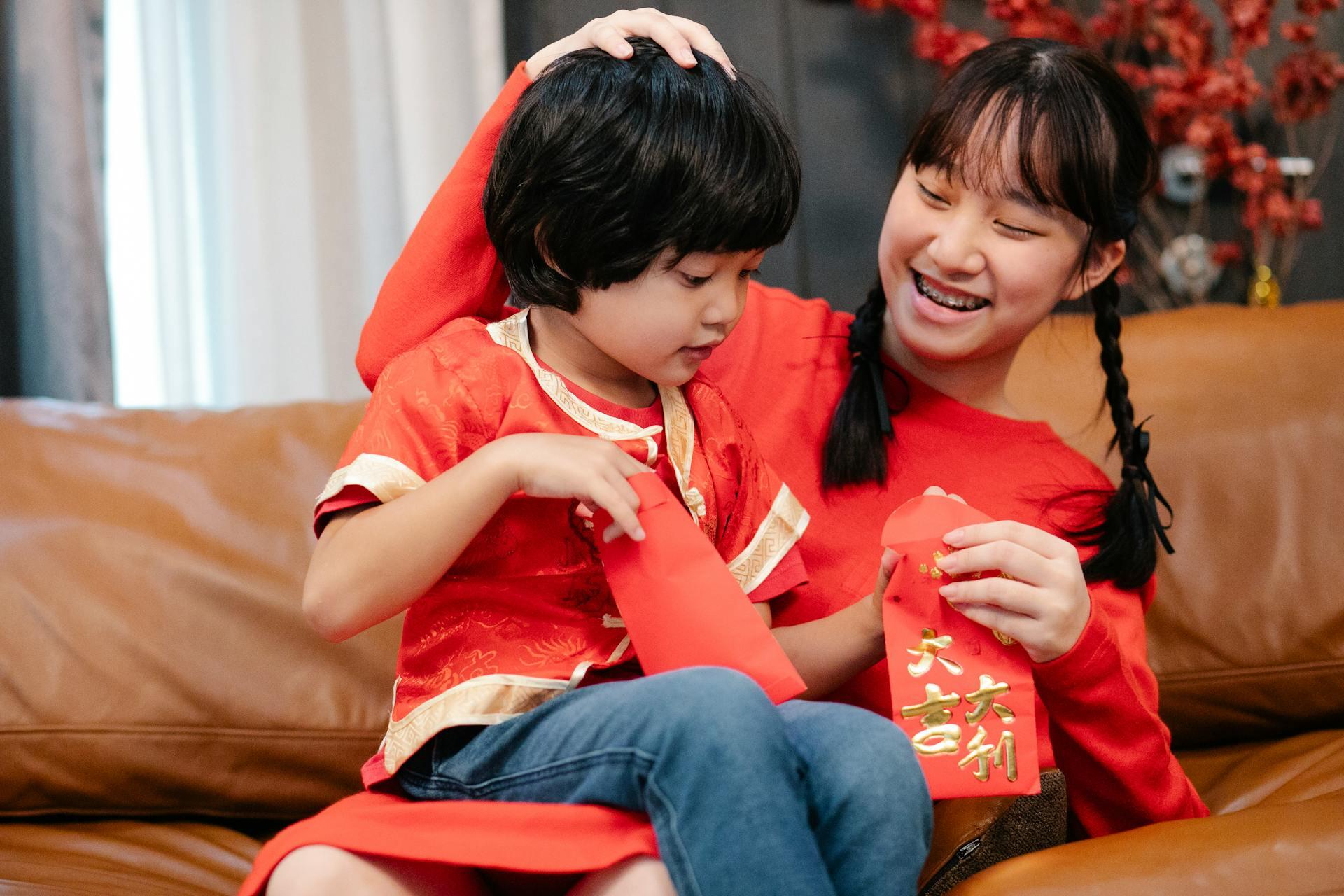 A young girl joyfully embraces a child, both wearing red traditional attire. They hold red envelopes, smiling warmly on a brown sofa, conveying festivity and joy.