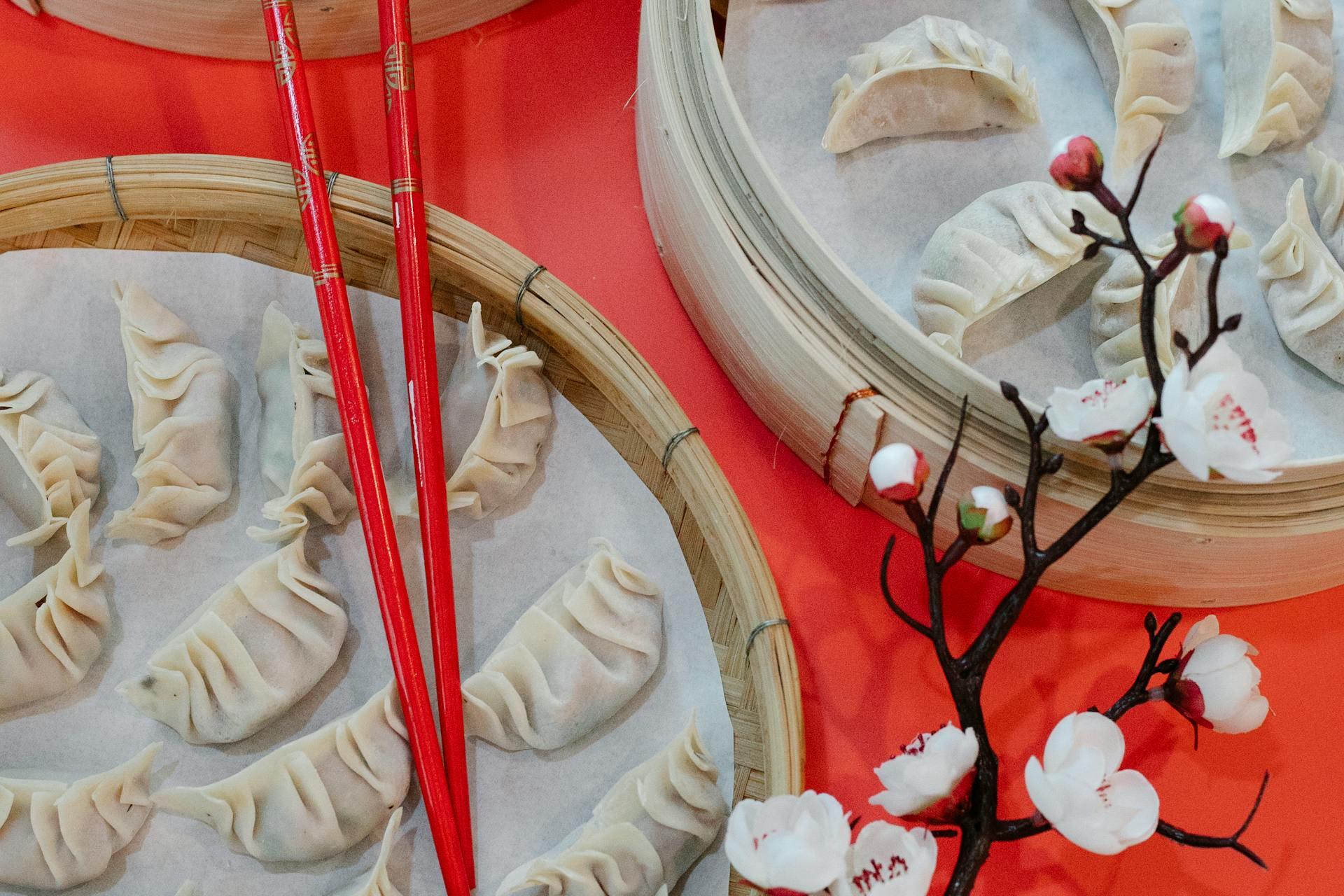 Steamed dumplings in bamboo baskets with red chopsticks rest on a vibrant red table. A branch of cherry blossoms adds an elegant touch.