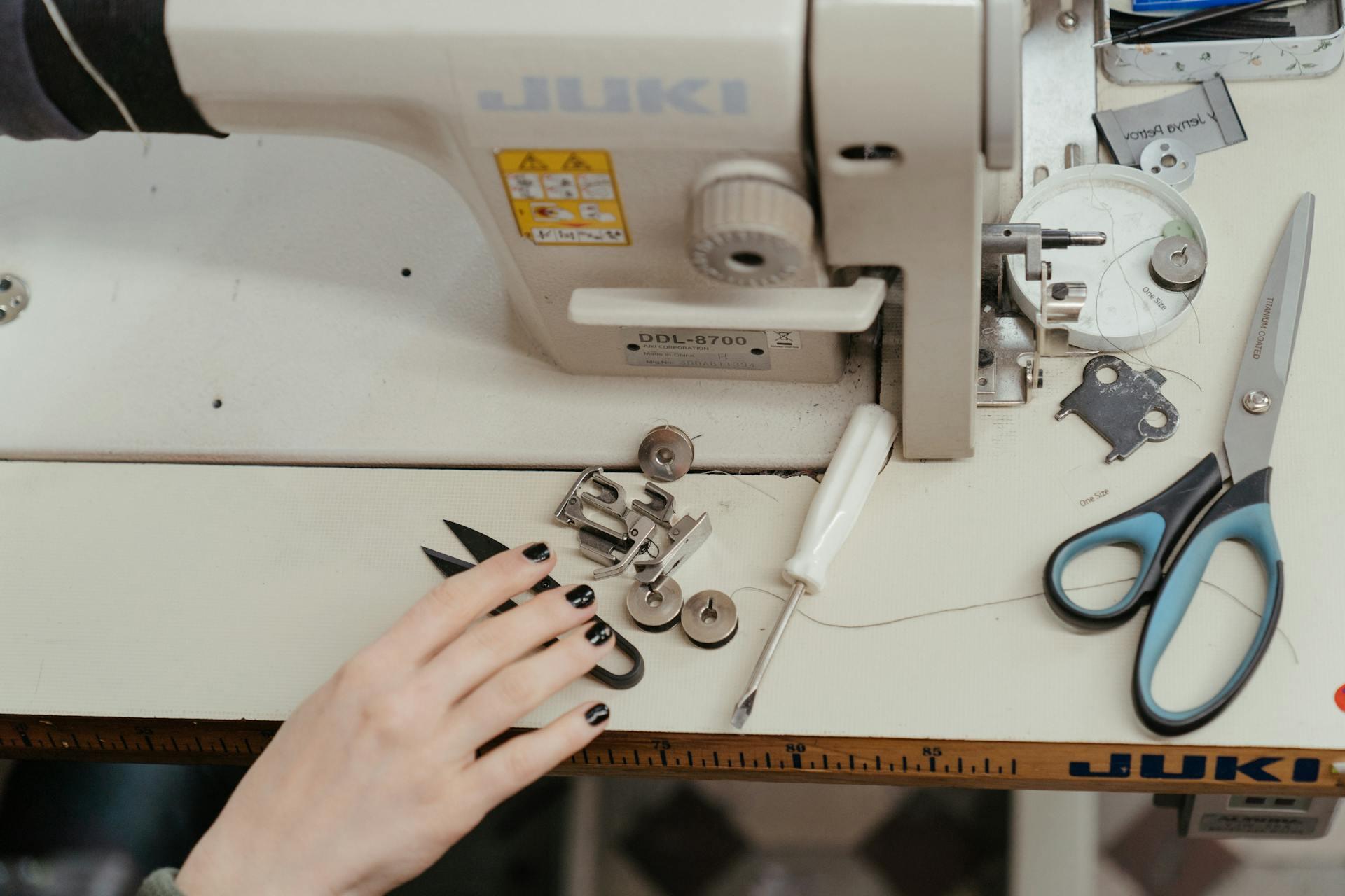 A hand with black nail polish arranges sewing machine parts on a Juki machine table. Scissors and tools are nearby, conveying a focused and creative workspace.