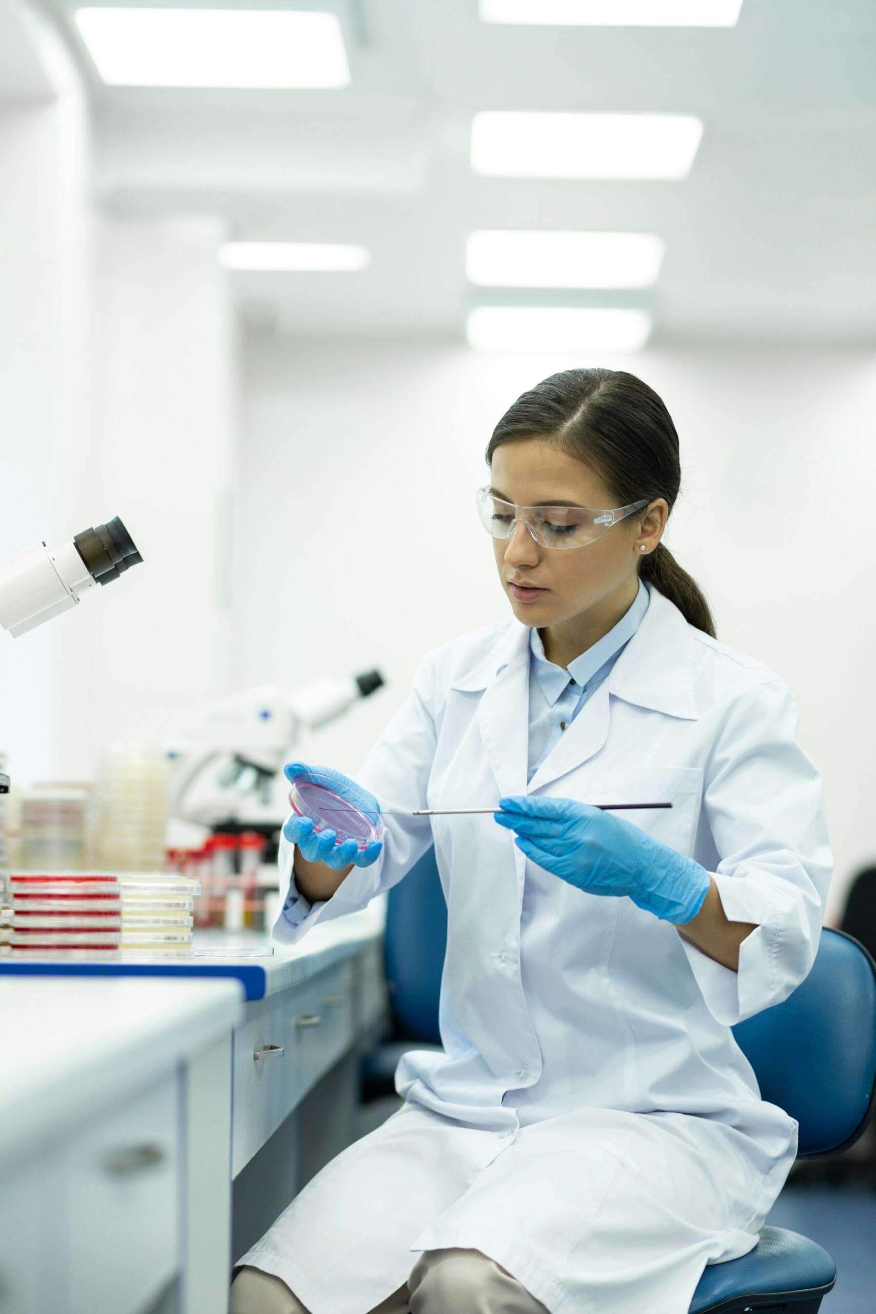 A person wearing a lab coat and goggles sitting at a lab bench.